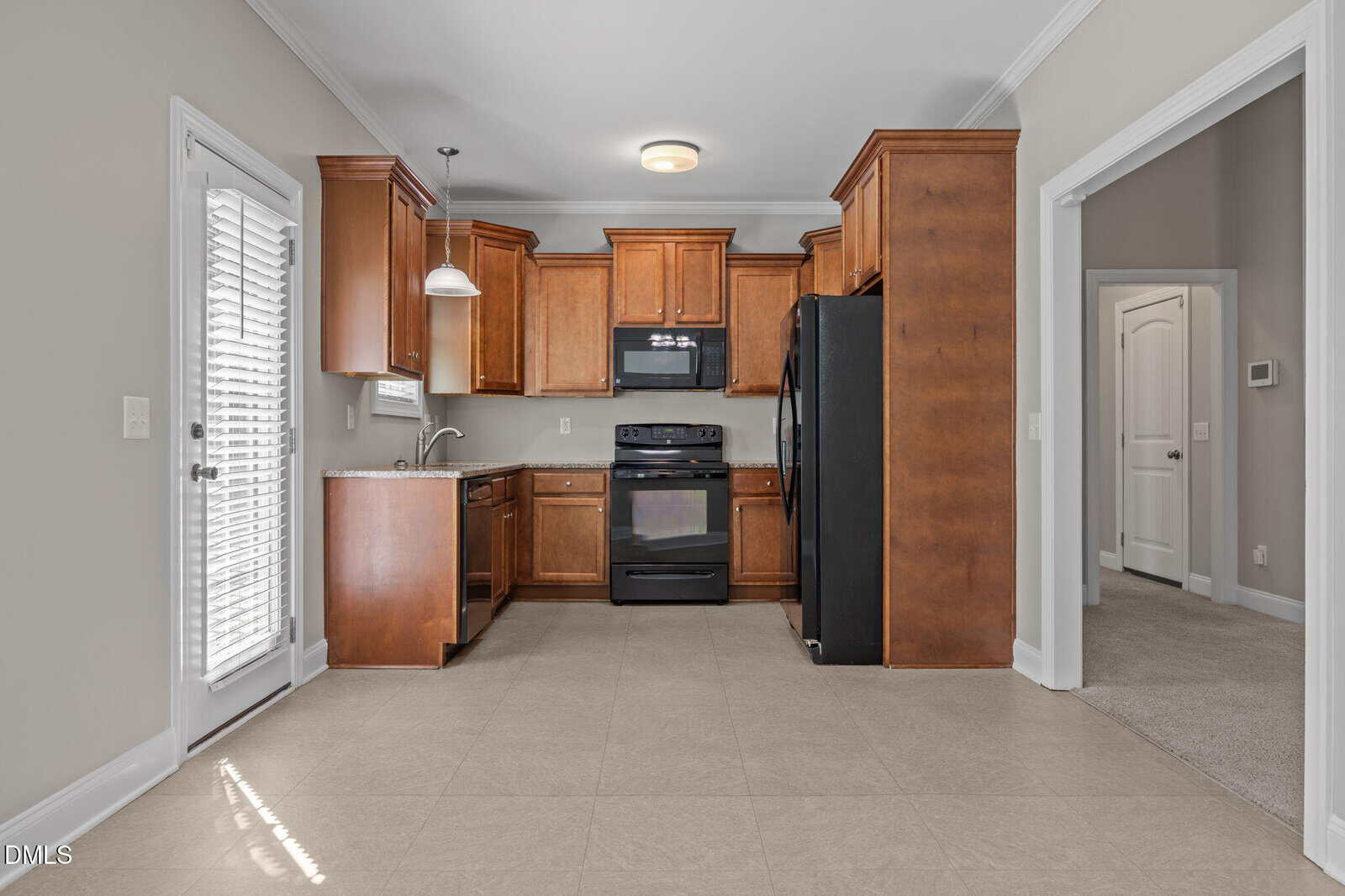 Modern kitchen with wooden cabinets, black appliances including a stove and refrigerator, beige tile floor, pendant lighting, and an adjacent carpeted room visible through an open doorway.