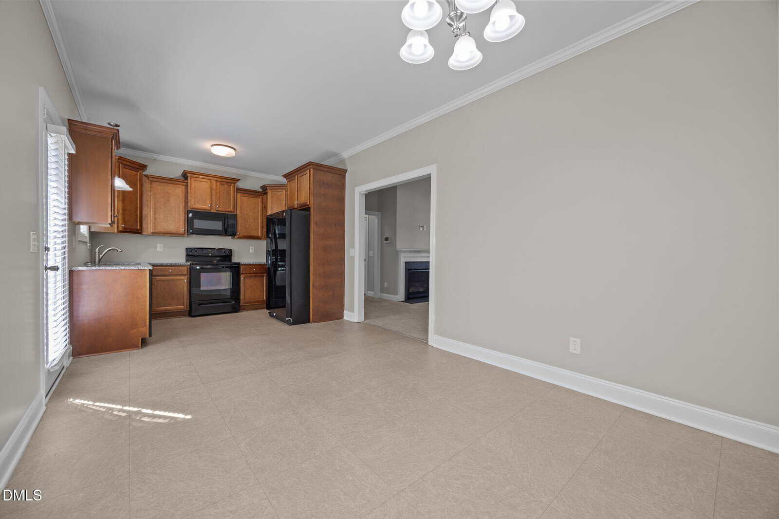 Spacious kitchen with wooden cabinets, black appliances, and beige tile flooring. Adjacent dining area with chandelier and large doorway leading to another room. Neutral wall colors and ample natural light.