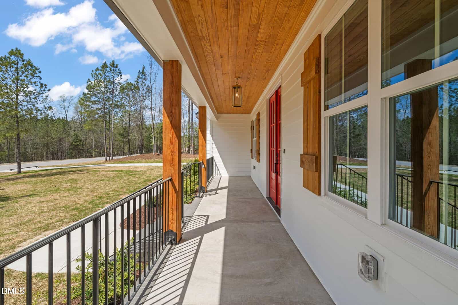 A front porch with a wooden ceiling, red front door, white siding, wooden window shutters, and black railing. The porch overlooks a grassy yard with trees and a clear blue sky in the background.