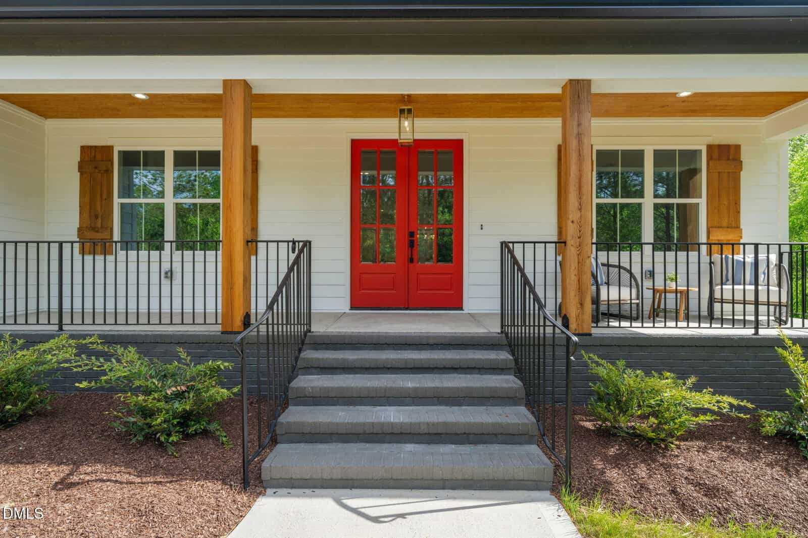 A modern house entrance with a bright red double front door, covered porch, black railings, wooden columns, white siding, and two outdoor seating areas on either side. Shrubs line the walkway leading to the steps.