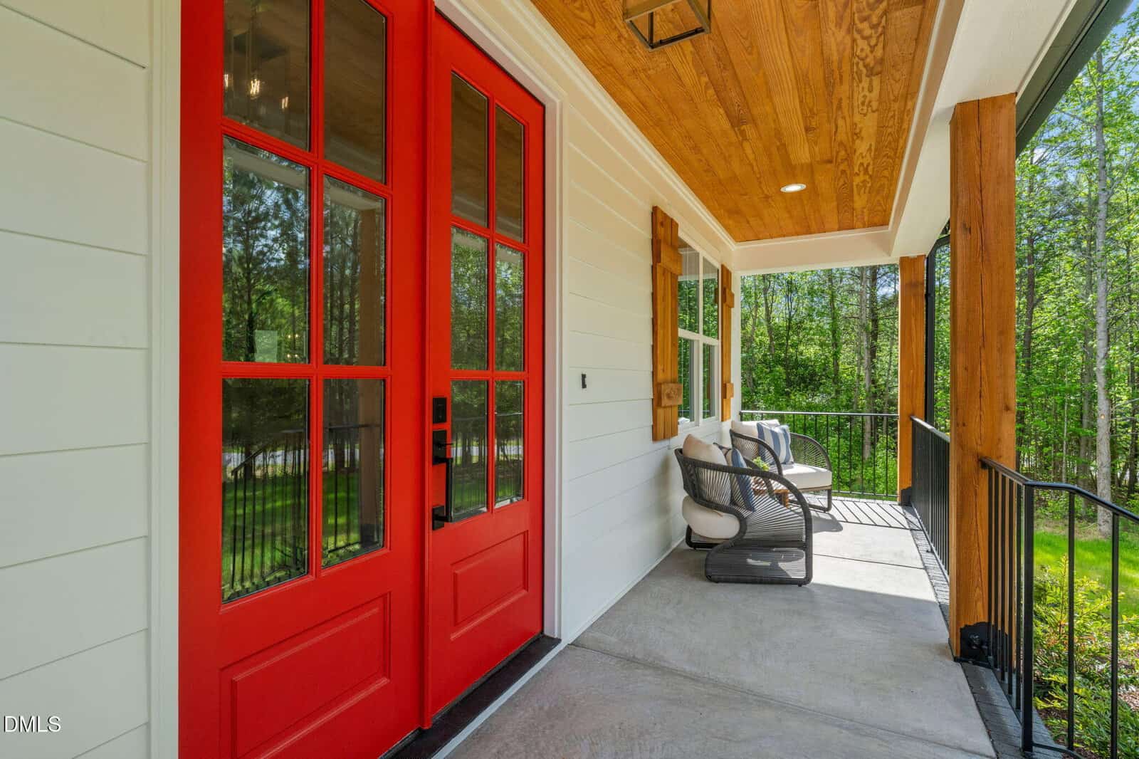 A cozy covered porch with red double doors, white siding, wood ceiling, two cushioned chairs, a small table, black railing, and views of green trees.