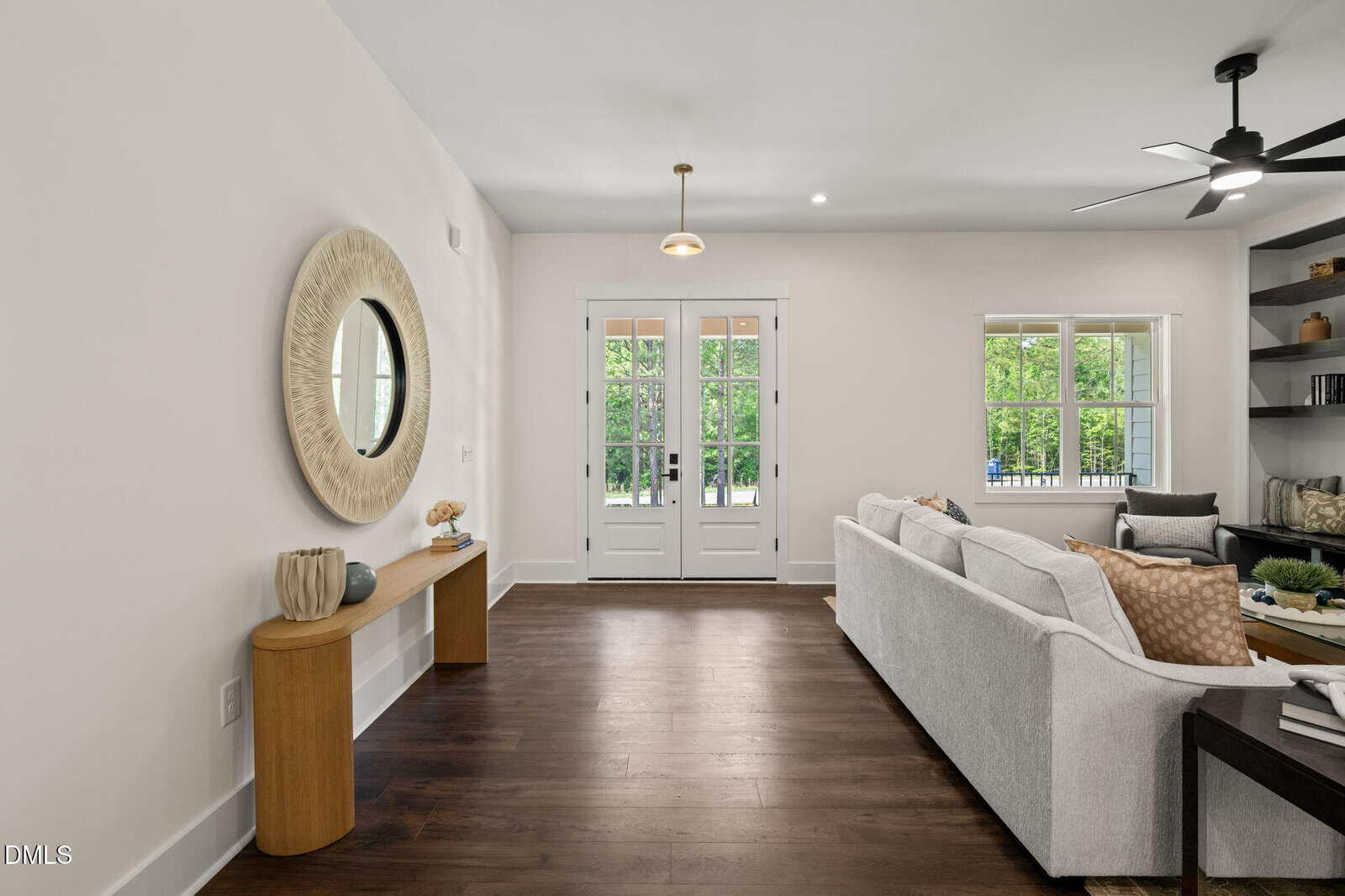 Modern living room with dark wood floors, a light gray sectional sofa, a wooden console table with decor, a round mirror, built-in shelves, large windows, and double doors opening to a green outdoor view.