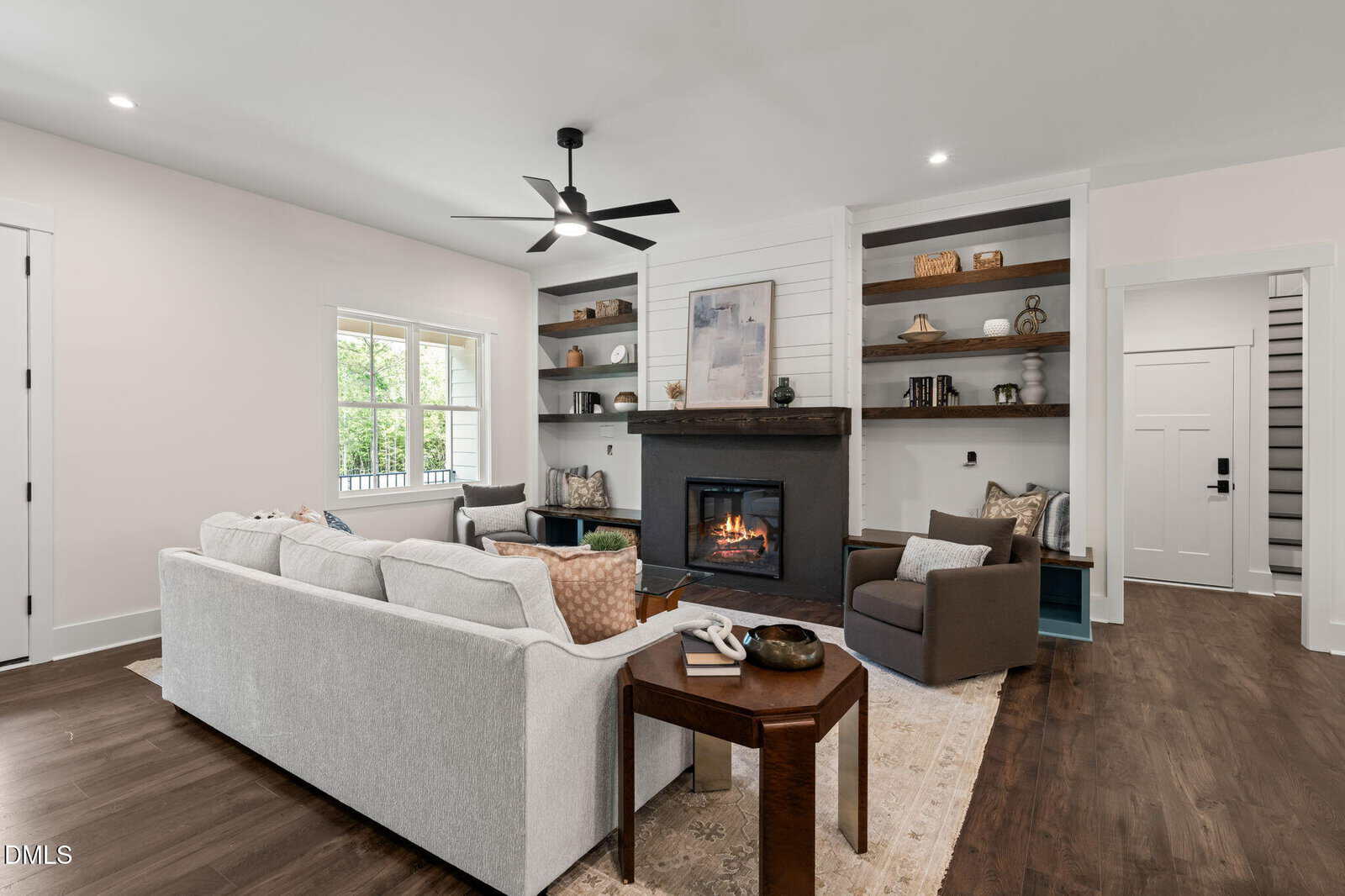 Modern living room with a light-colored sofa, armchair, wooden coffee table, fireplace, built-in shelves, and large window. The room has dark wood floors and white walls, creating a cozy and inviting space.