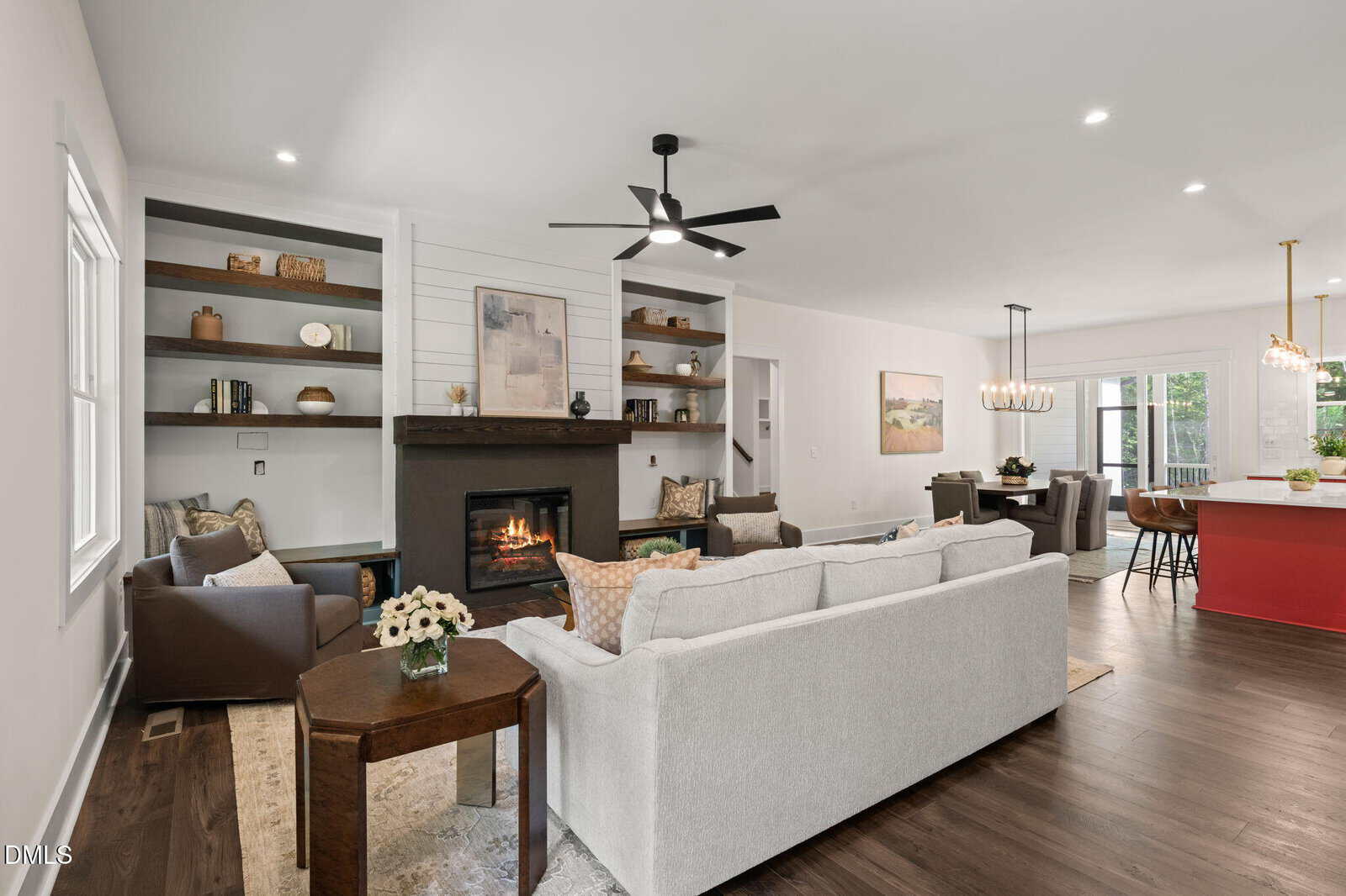 Modern living room with a gray couch, armchairs, a fireplace, built-in shelves, and a ceiling fan. Open layout leads to kitchen with red island and dining area. Large windows provide natural light.