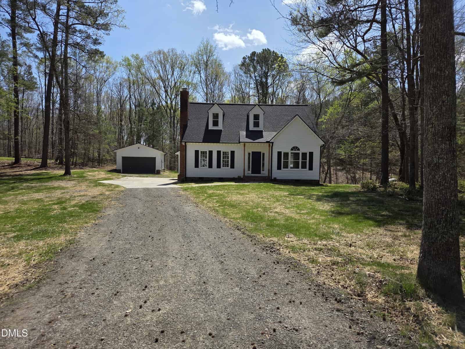 A two-story white house with dark shutters and a front porch sits at the end of a gravel driveway, surrounded by trees and grass. A detached garage is visible to the left of the house.