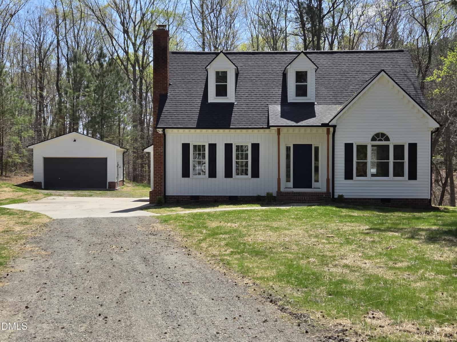 A white two-story house with dark roof shingles, a central chimney, and black shutters. A detached two-car garage sits to the left, connected by a gravel driveway. Trees and grass surround the property.