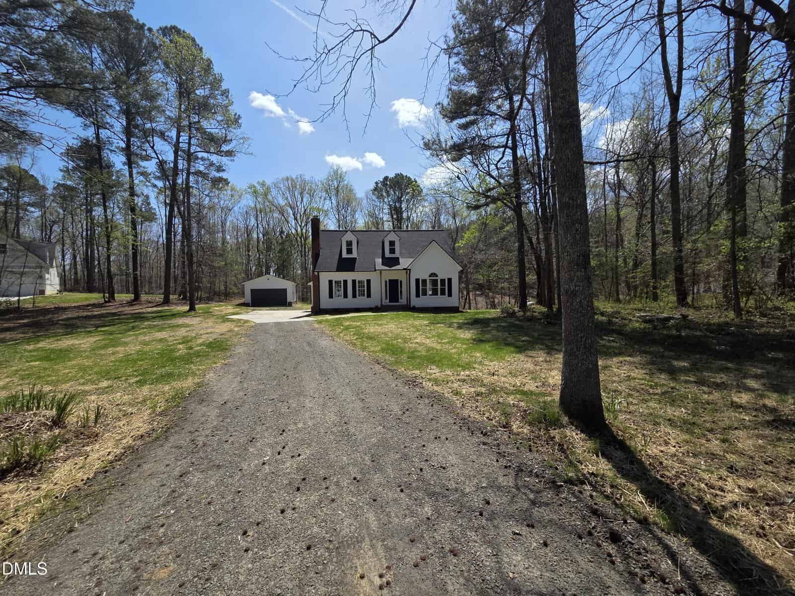 A two-story white house with dormer windows and a detached garage sits at the end of a long gravel driveway, surrounded by tall trees and a grassy yard under a partly cloudy sky.