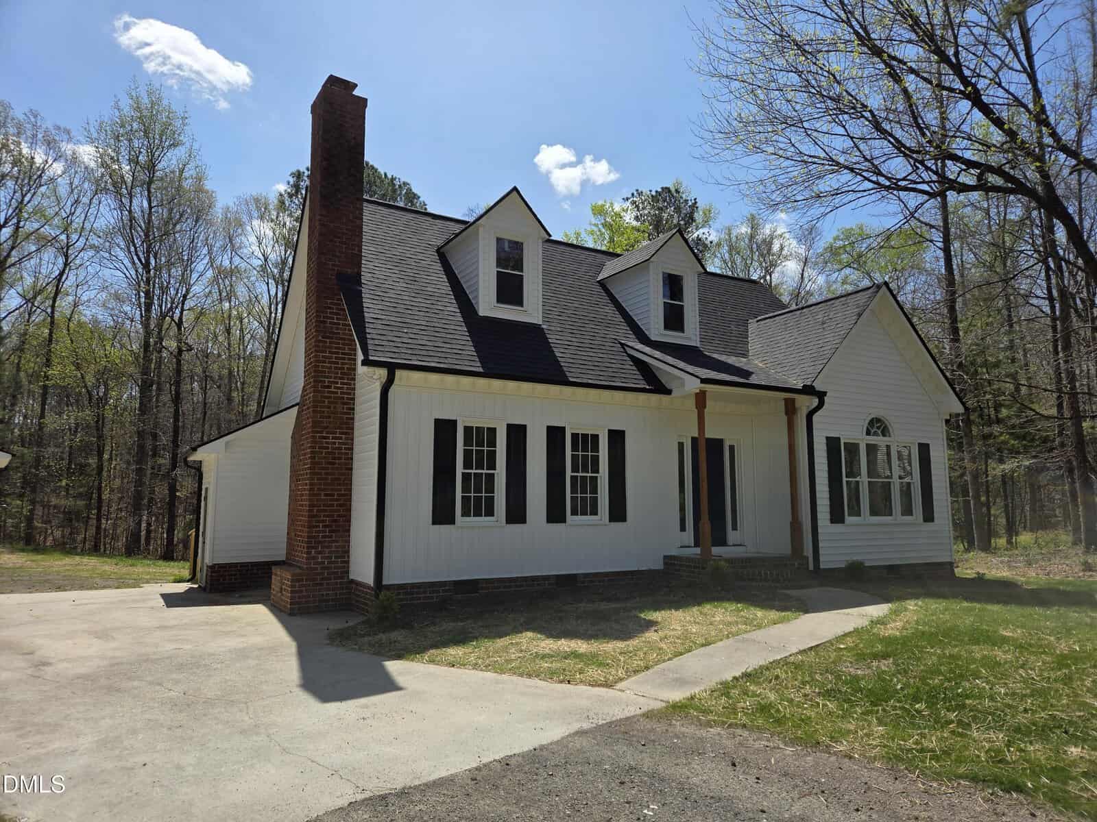 A white house with black shutters and a steep roof, featuring a prominent brick chimney, two dormer windows, a small front porch with columns, and a driveway surrounded by trees and grass.