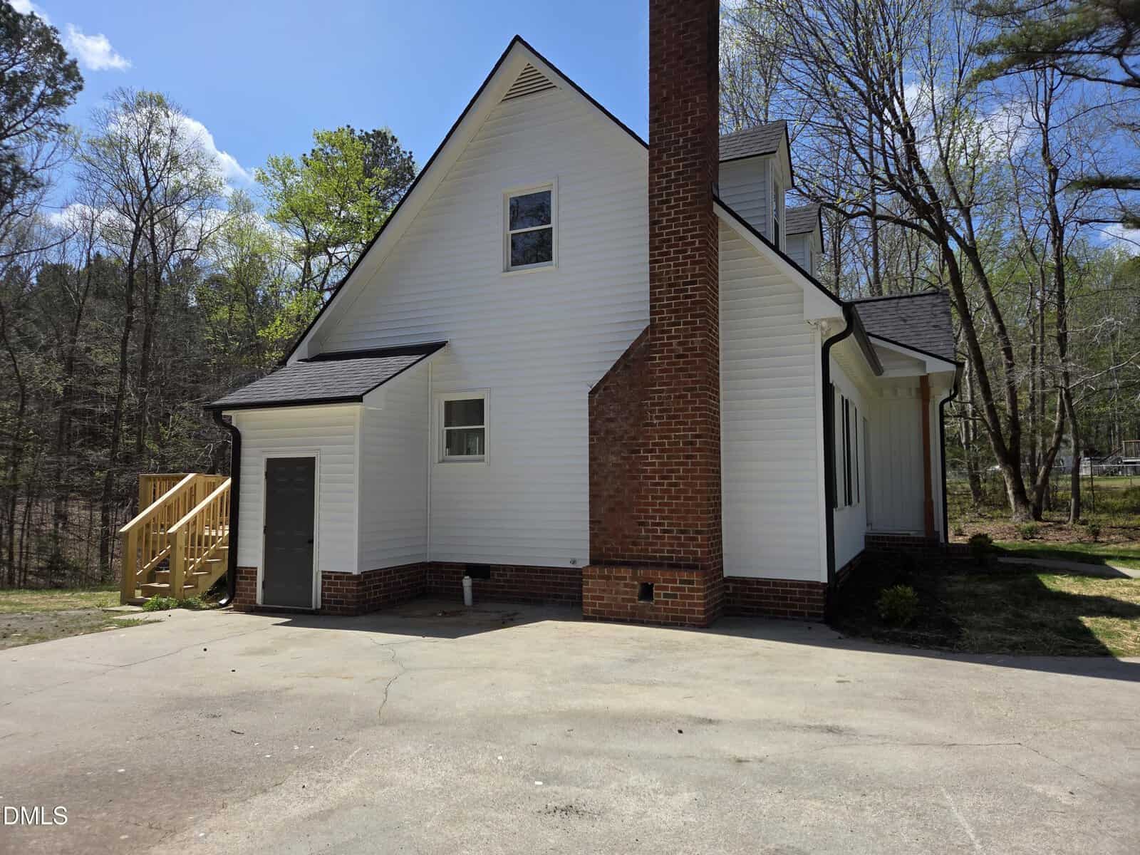 A two-story white house with black trim, a large red brick chimney, a gray side door, and wooden stairs leading to a separate entrance. The house is surrounded by trees and has a concrete driveway in front.