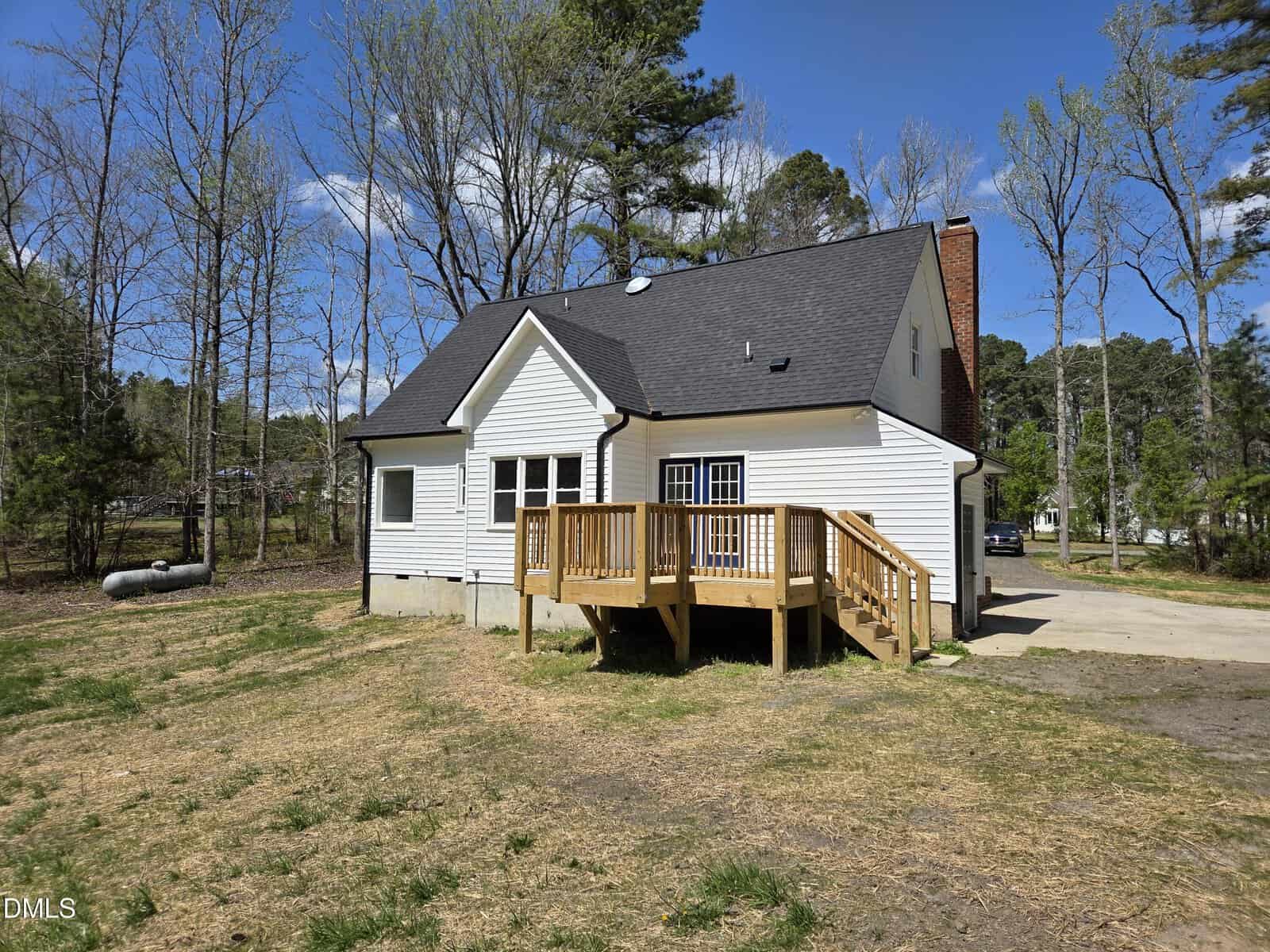 A white house with black roof sits among trees. A small wooden deck with stairs leads to a grassy yard. A propane tank is visible on the left, and a driveway is on the right.