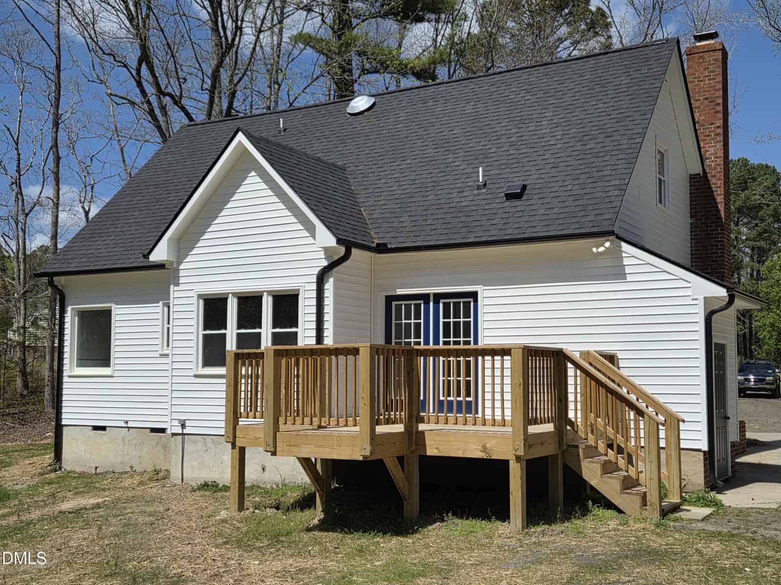 White house with black roof, chimney, and multiple windows, featuring a wooden deck with stairs leading to a grassy yard. Trees without leaves surround the home under a sunny blue sky.