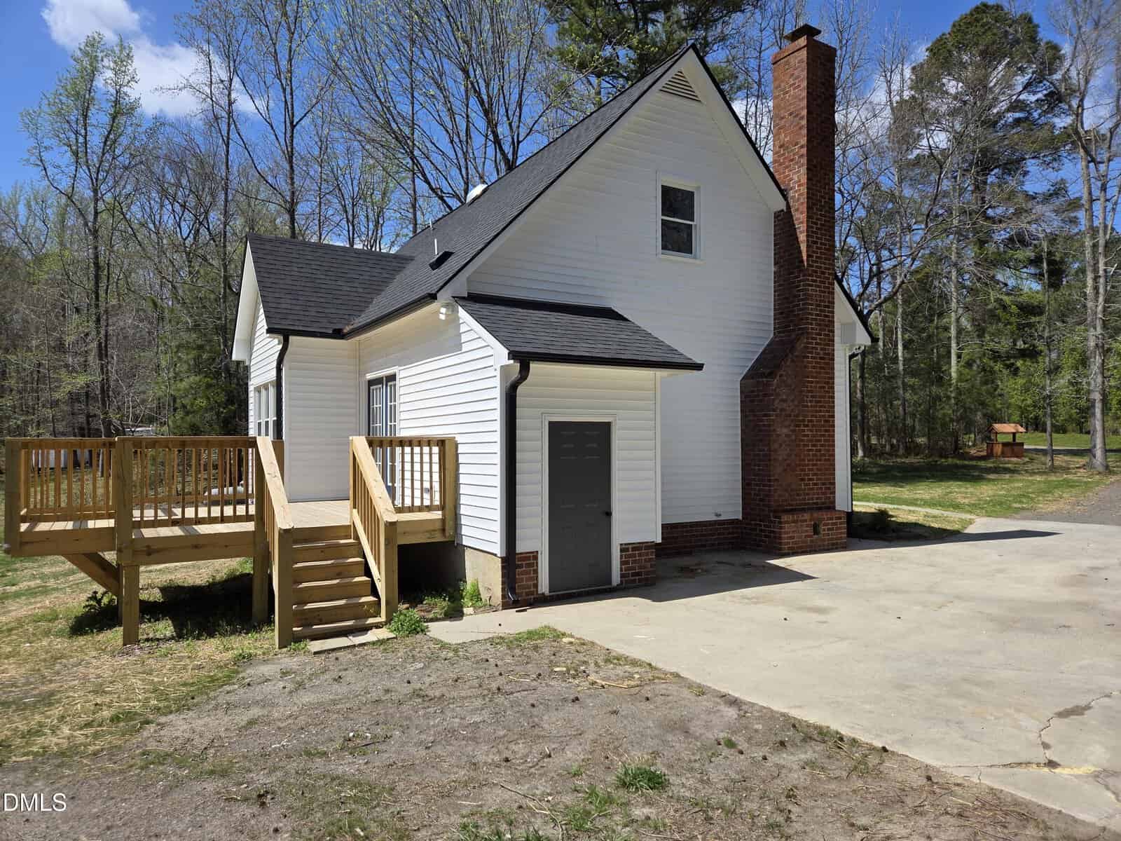 A white two-story house with black roof, large brick chimney, wooden deck with stairs, and concrete driveway, surrounded by trees and a grassy yard on a sunny day.