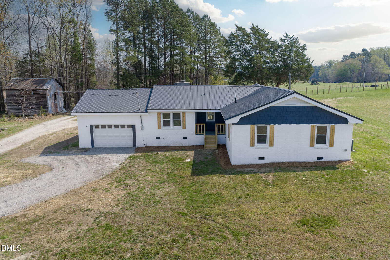 A white ranch-style house with a dark metal roof and attached garage, surrounded by grass and trees, sits on a rural property under a partly cloudy sky.