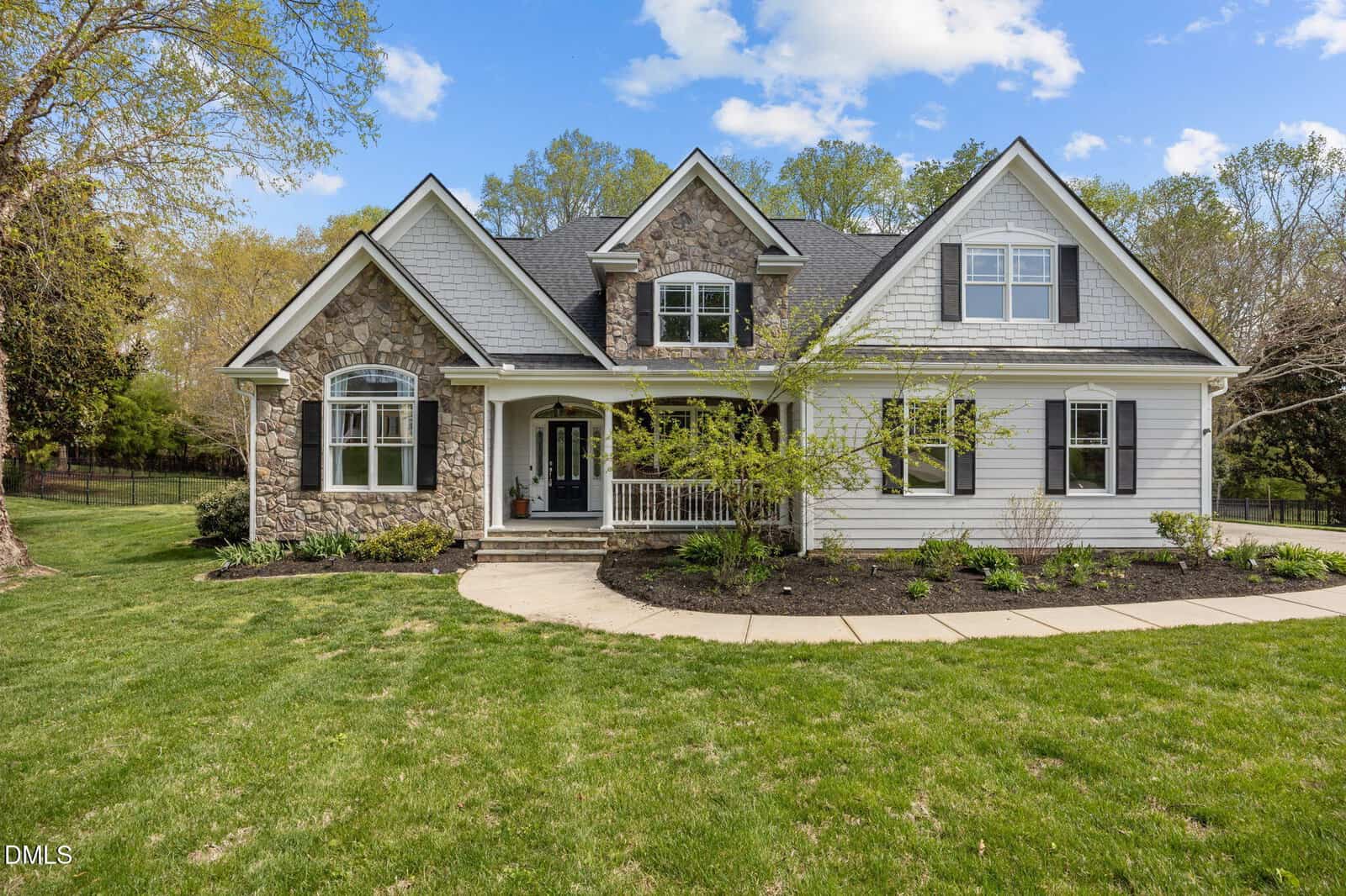 A two-story suburban house with stone and white siding exterior, a covered front porch, dark roof, and landscaped yard with young trees and shrubs on a sunny day.
