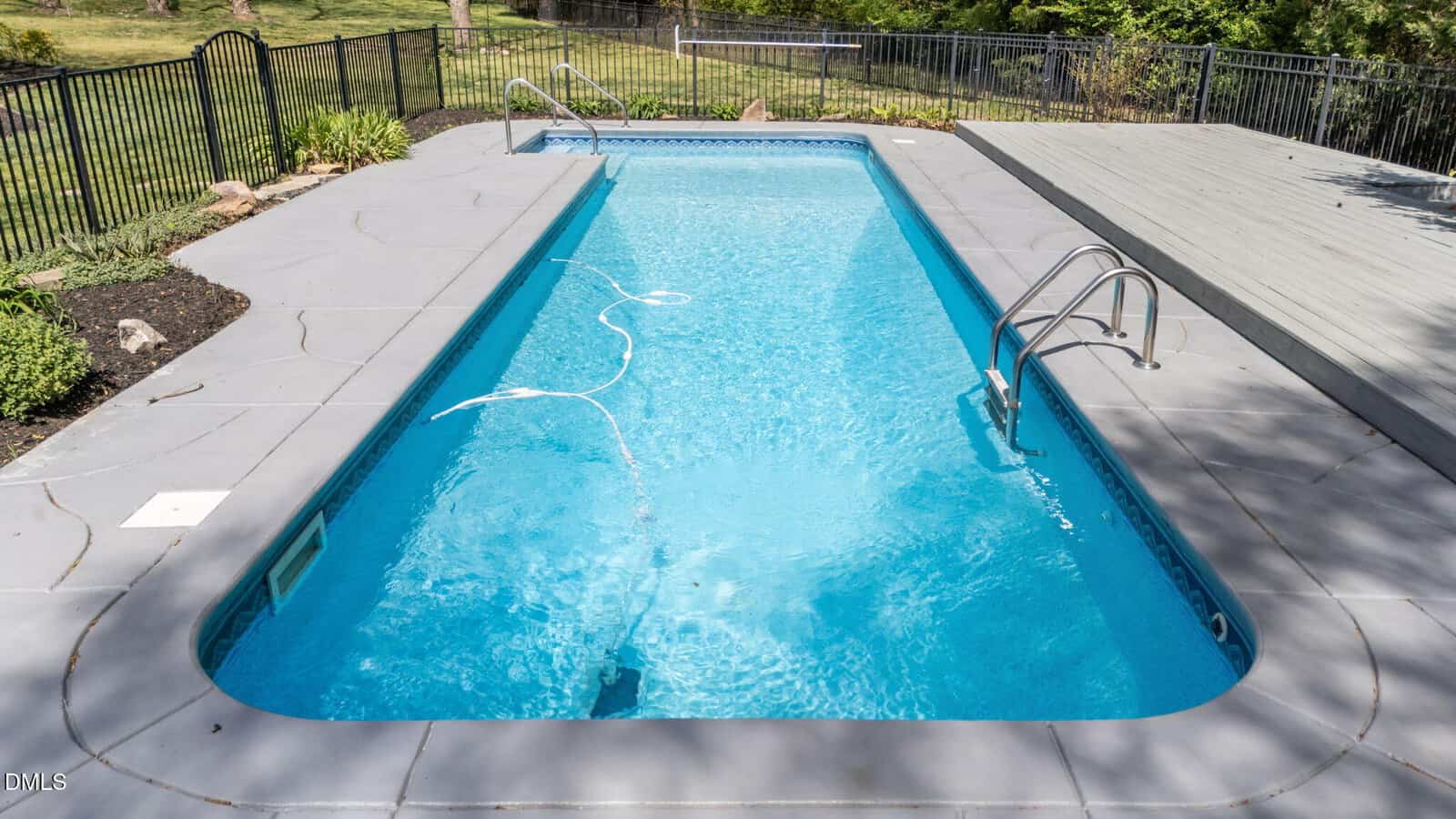 Rectangular in-ground swimming pool with blue water, surrounded by gray concrete decking, metal pool ladder, and a fenced grassy yard in the background. A white pool vacuum hose floats on the surface.