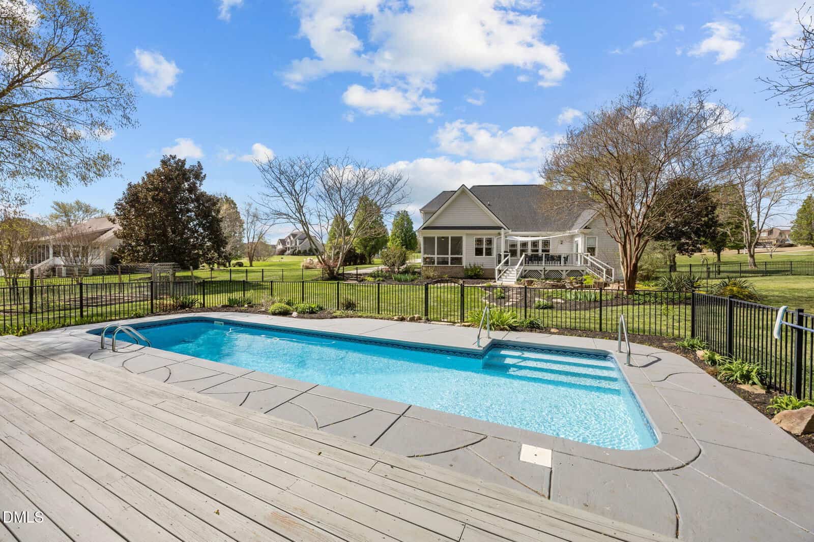 Rectangular backyard pool with handrails, surrounded by a black fence, wooden deck, green lawn, trees, and a white house in the background under a blue sky with clouds.
