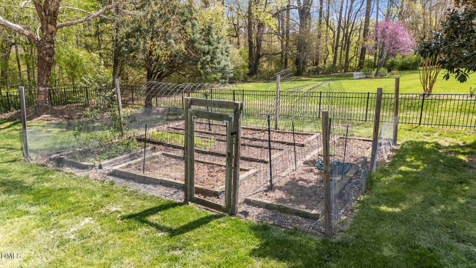 A fenced garden plot with raised beds sits on a grassy lawn, surrounded by trees and a black metal fence in the background under a sunny sky.