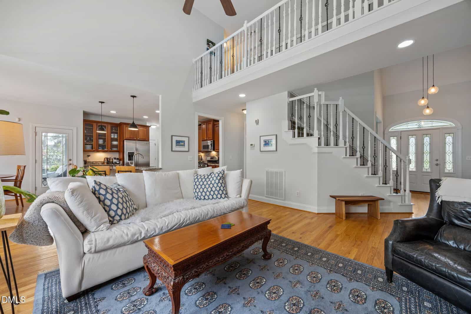 Spacious living room with white sofa, wooden coffee table, and blue patterned rug. Open layout connects to kitchen with wood cabinets. Staircase and balcony visible, along with large windows and hardwood floors.