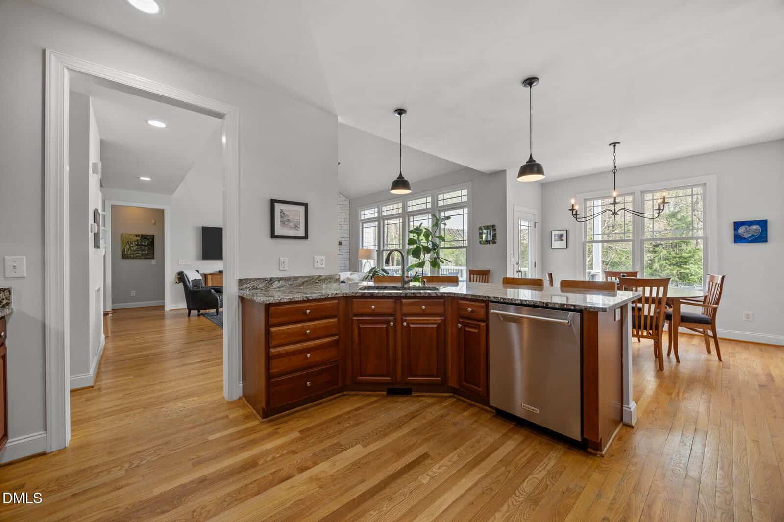 A spacious kitchen with wooden floors, dark wood cabinets, granite countertops, stainless steel dishwasher, and large windows offering natural light. The dining area is visible in the background with a chandelier above the table.
