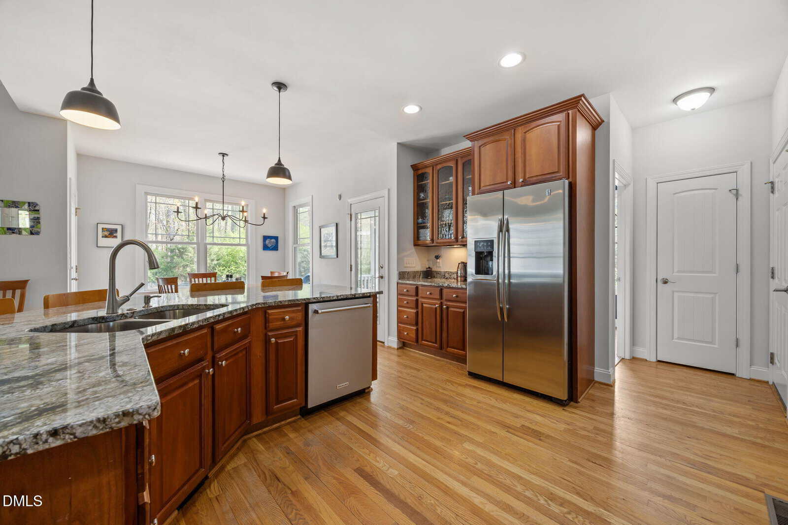 A spacious kitchen with wood cabinets, stainless steel appliances, granite countertops, and an island with a sink. Hardwood floors extend into a dining area with large windows and pendant lights overhead.