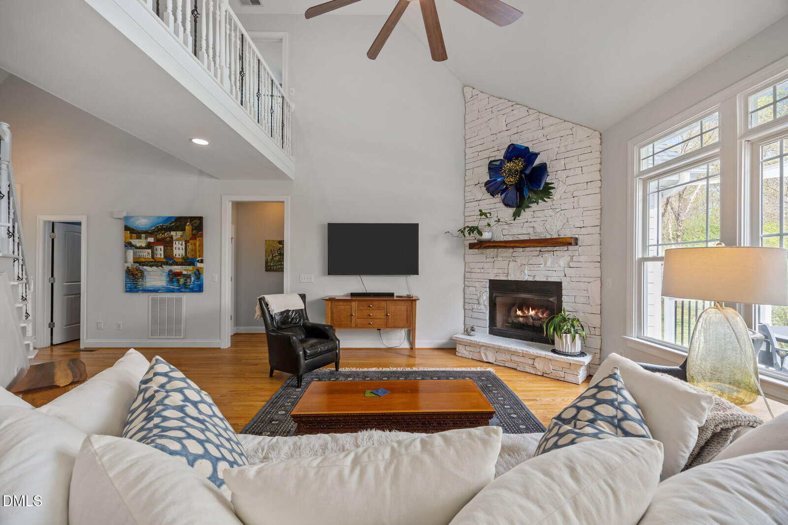 A living room with a white stone fireplace, a mounted television, a wooden coffee table, an armchair, a sofa with patterned pillows, and large windows allowing natural light into the space.
