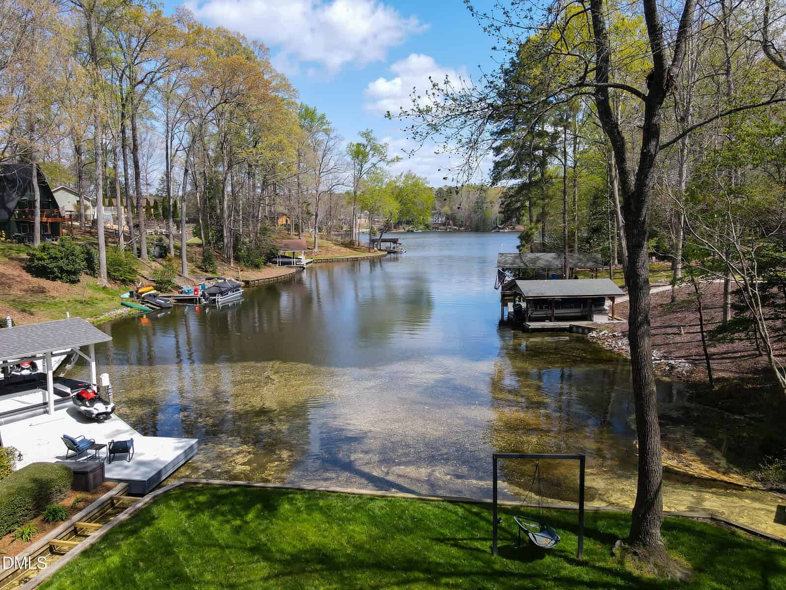 A tranquil lake bordered by trees and houses, with docks and boats along the shoreline. The foreground features a grassy yard with a hammock beside the water, under a partly cloudy blue sky.