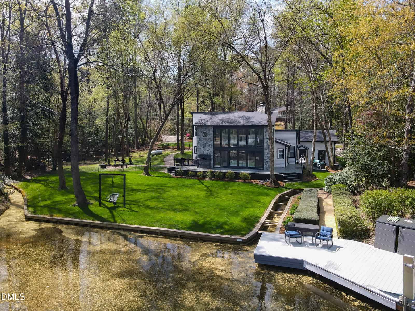 Modern lakeside house with large glass windows surrounded by tall trees, a green lawn, a swing, and a white deck with chairs next to clear water. Pathways lead from the house to the deck and around the yard.
