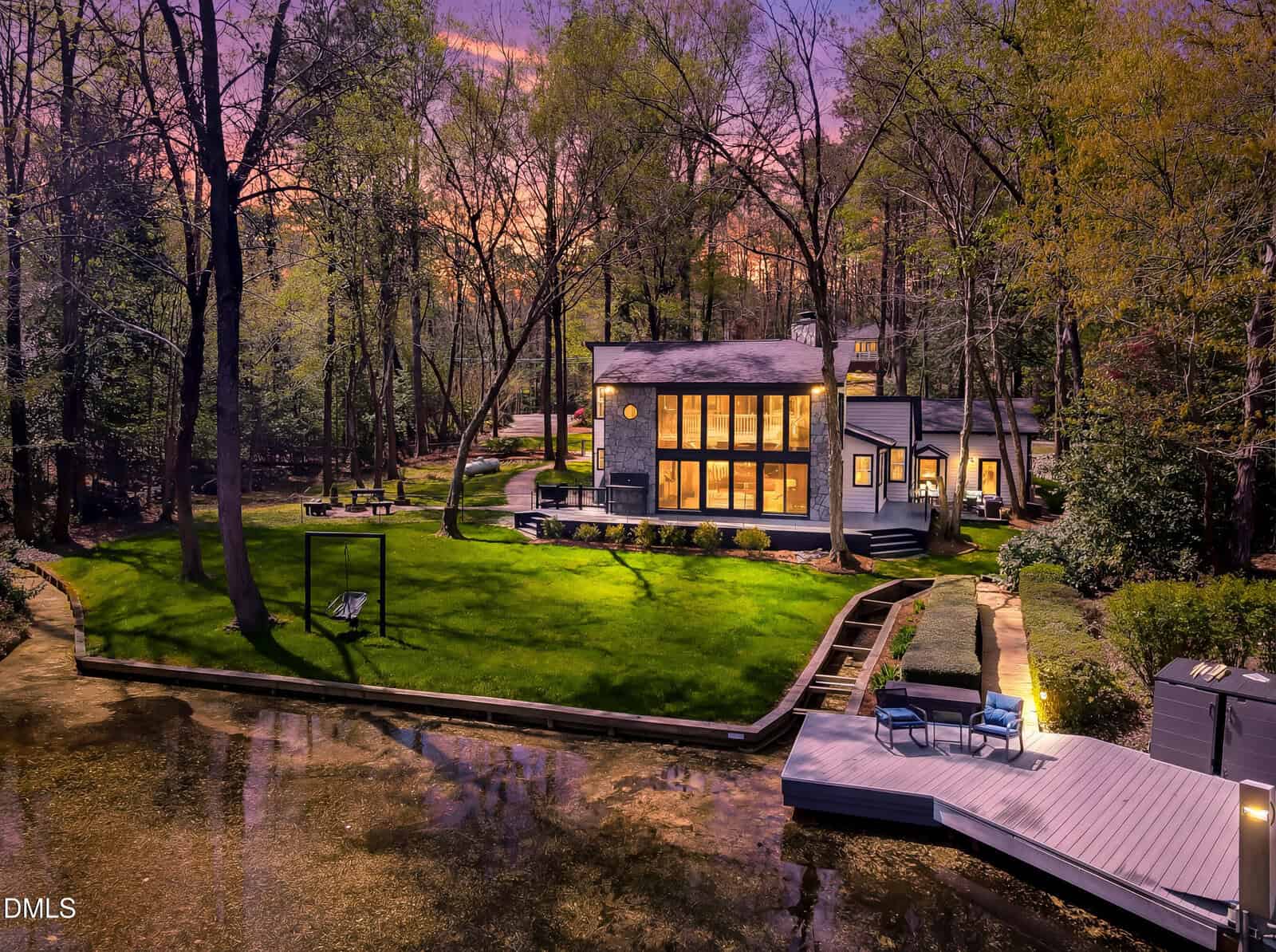 Modern house with large windows illuminated at dusk, surrounded by tall trees and lush lawn, next to a calm body of water with a dock, outdoor seating, and a swing set. The sky is a mix of purple and orange hues.