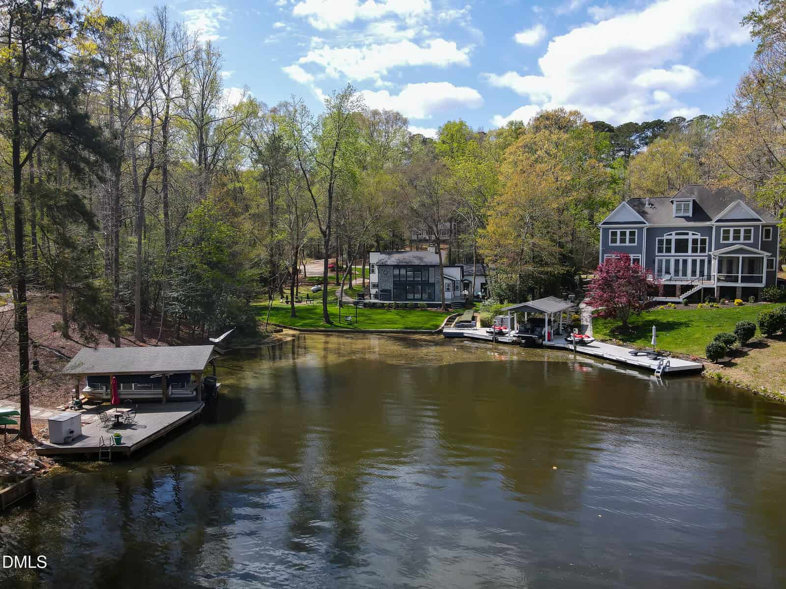 A calm lake bordered by trees with two docked boats and waterfront houses; one house is large with gray siding and a deck, another is smaller with large windows. The sky is partly cloudy.