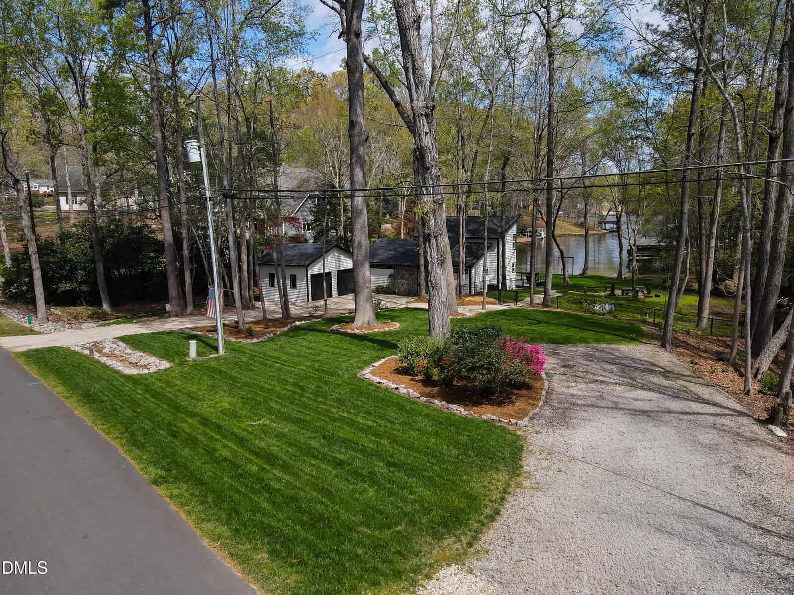 A house with a gray roof sits among tall trees beside a lake. The yard features green grass, flower beds, and a gravel driveway. A paved road runs in front, and power lines cross overhead.