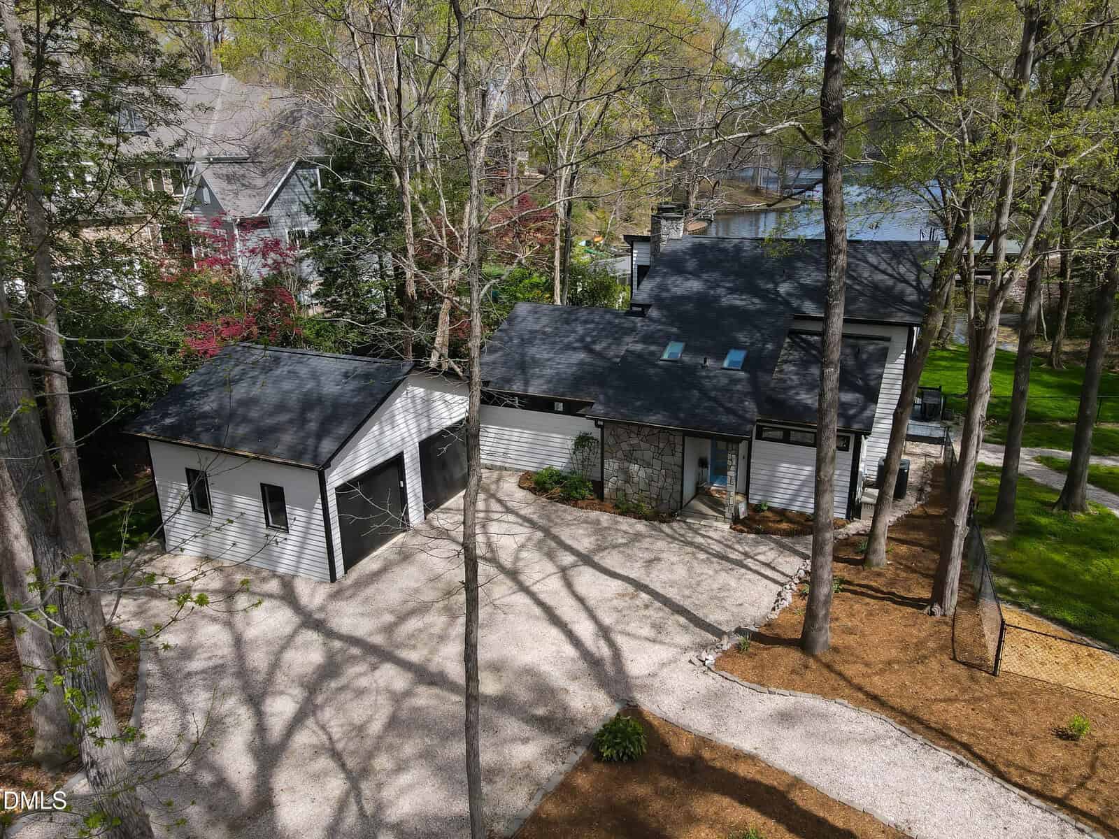 A modern house with a detached garage sits among tall trees, surrounded by a gravel driveway and landscaped beds. The home features dark-framed windows and light-colored siding, with a lake visible in the background.
