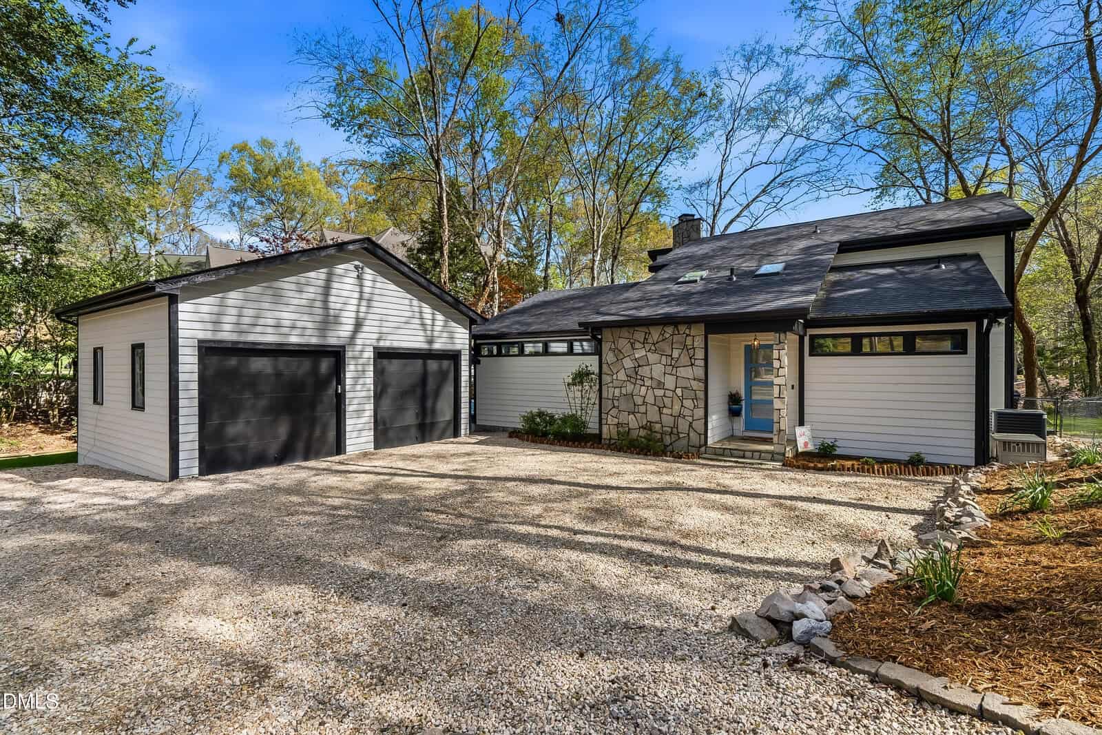 Modern house with stone and gray siding exterior, black garage doors, gravel driveway, and landscaped yard with trees and mulch beds under a clear blue sky.