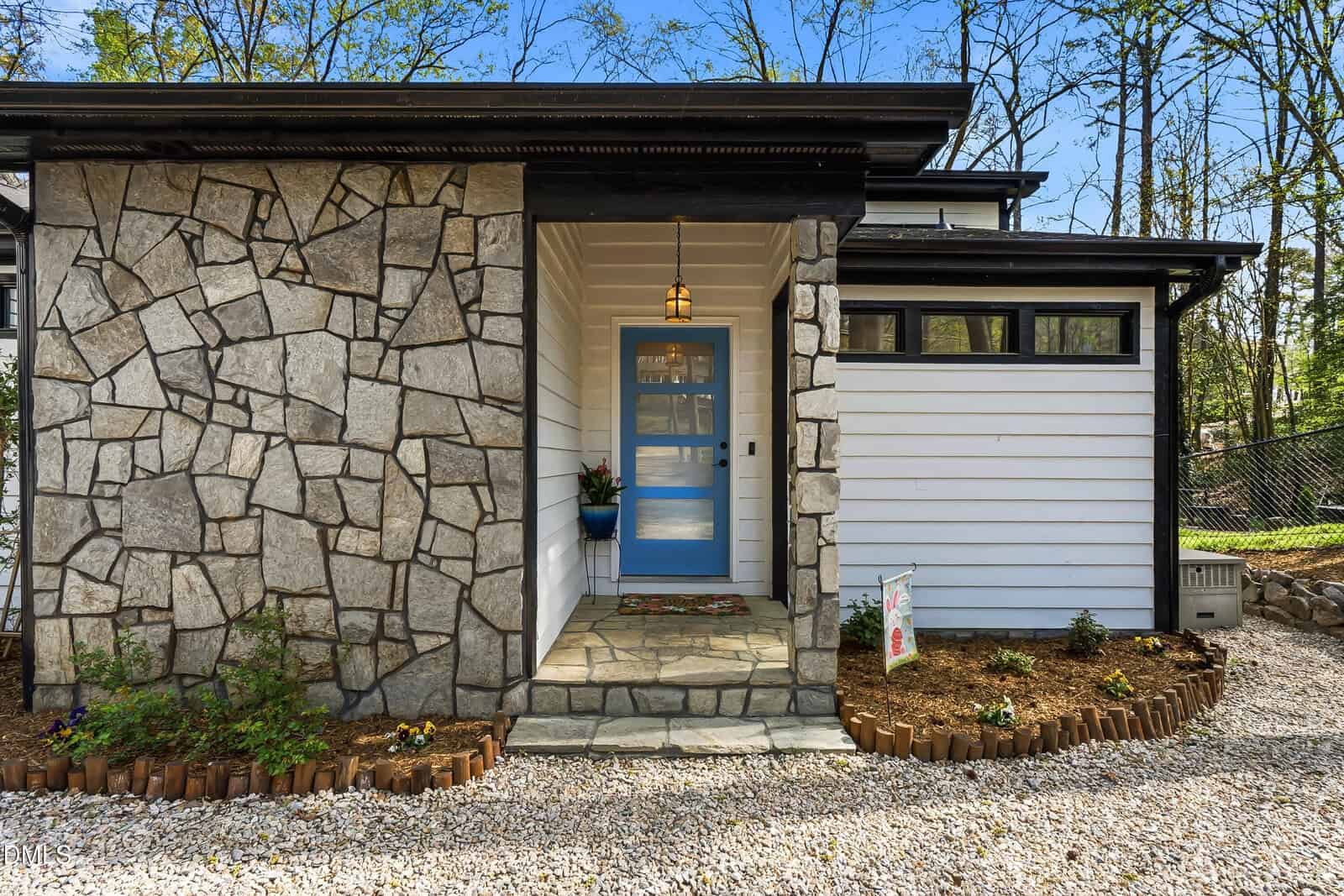 A modern house exterior with a stone and white wood facade, blue front door, small covered porch with a hanging light, potted plant, garden beds, and gravel walkway, surrounded by trees and greenery.