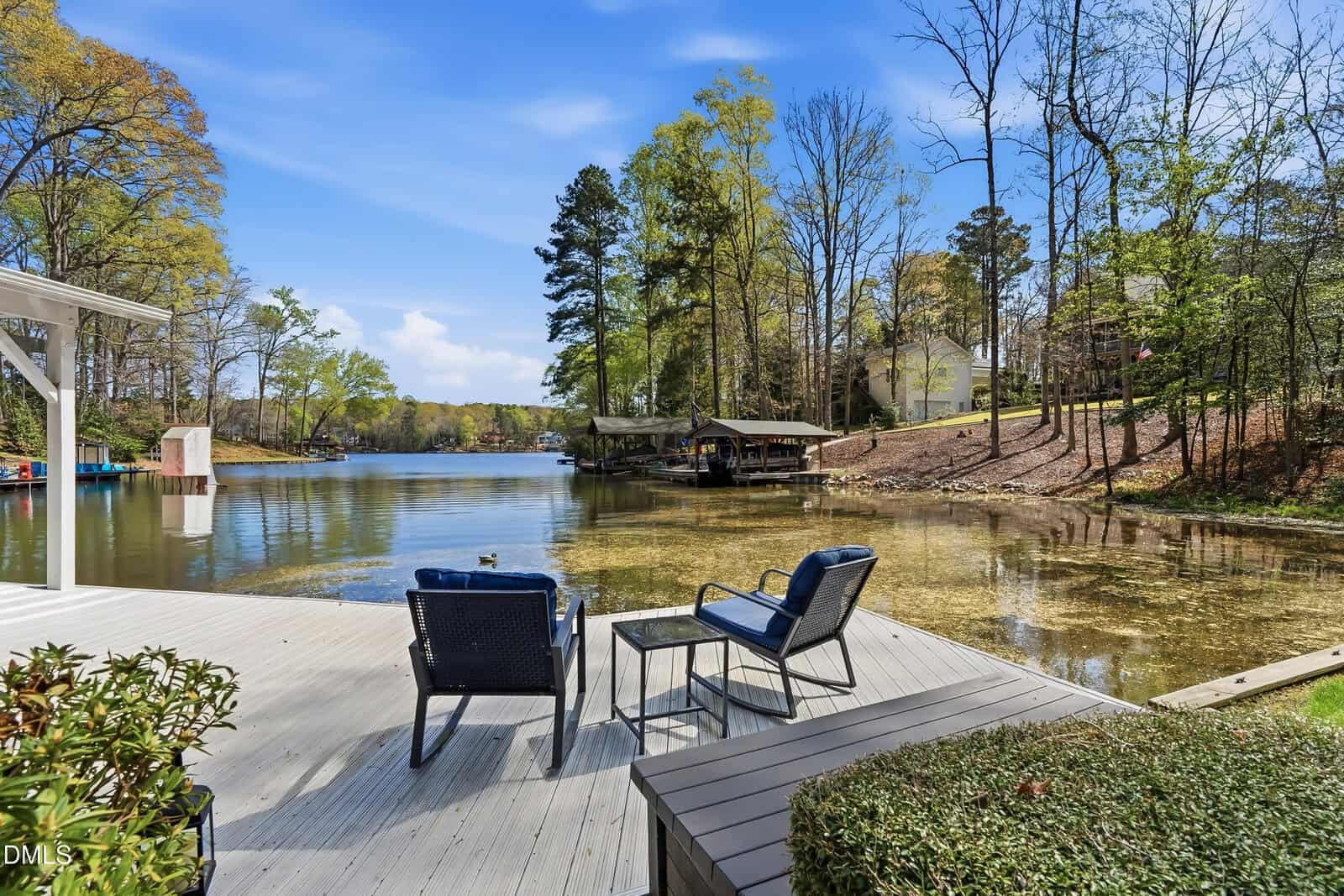 Two chairs and a small table sit on a lakeside wooden deck, overlooking calm water, trees, and nearby houses under a blue sky with scattered clouds.