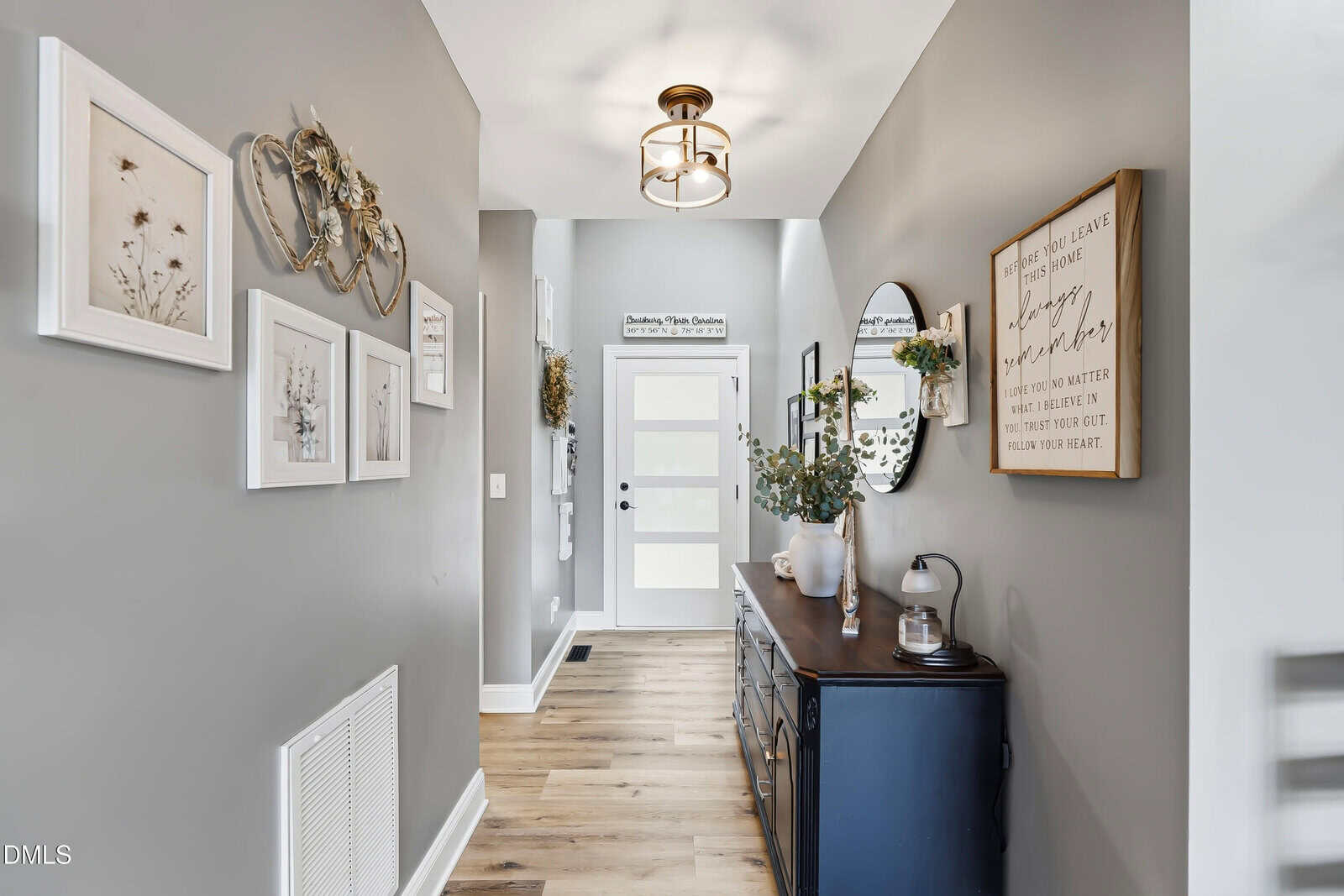 A modern hallway with gray walls, wooden floor, framed art, a round mirror, a black console table with decor, and a white door at the end. A gold light fixture hangs from the ceiling.