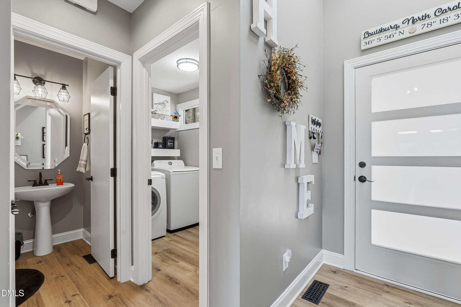 A modern entryway with light wood flooring, a white front door, wall decor reading “HOME,” and views into a laundry room and a bathroom with gray walls and white trim.