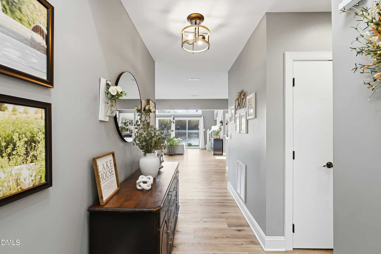 A bright hallway with light wood floors, gray walls, and white trim. The left side features a black console table decorated with plants, a round mirror, and framed art. Natural light streams in from windows at the far end.