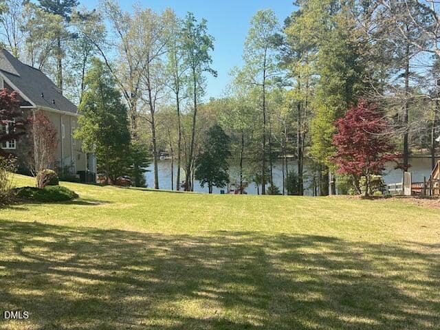 A grassy backyard slopes down toward a lake surrounded by tall trees. A house is partially visible on the left, and there are red and green-leaved trees scattered throughout the yard. The sky is clear and blue.
