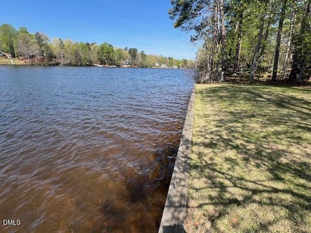 A calm lake bordered by a grassy area and tall trees, with blue sky above. There’s a wooden retaining wall separating the grass from the water. Houses and more trees are visible across the lake.