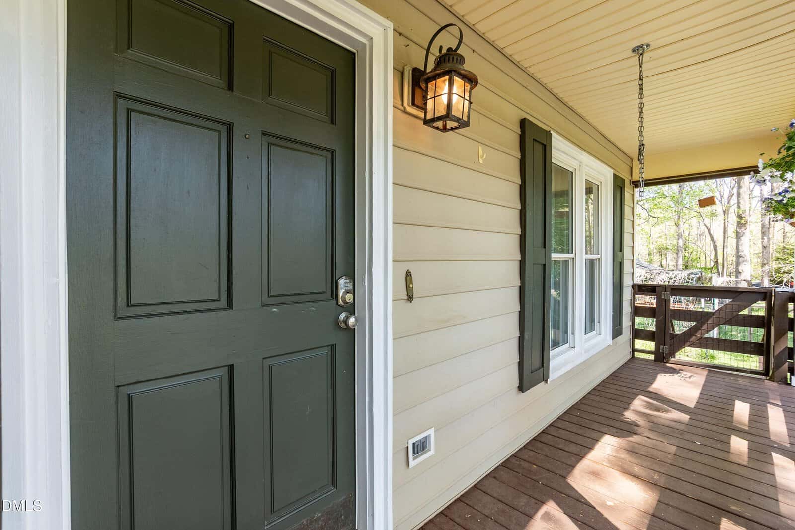 A green front door with a silver handle is next to two white-framed windows on a beige house. A black lantern-style light fixture and porch swing are attached to a wooden deck that overlooks a yard.