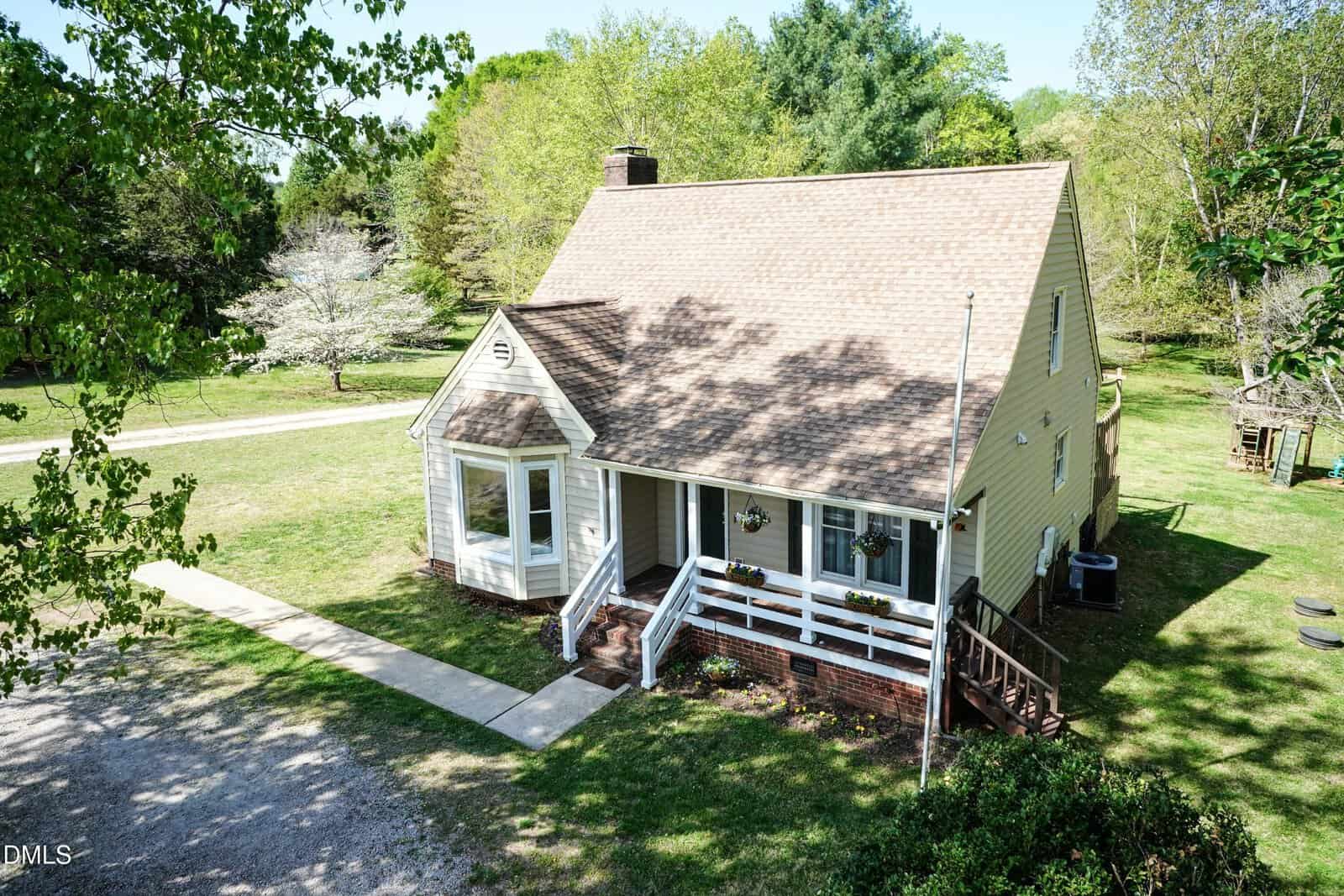 A light-colored, two-story house with a steep roof, white porch railing, and bay window sits on a grassy lot with trees in the background and a gravel driveway leading to the front steps.