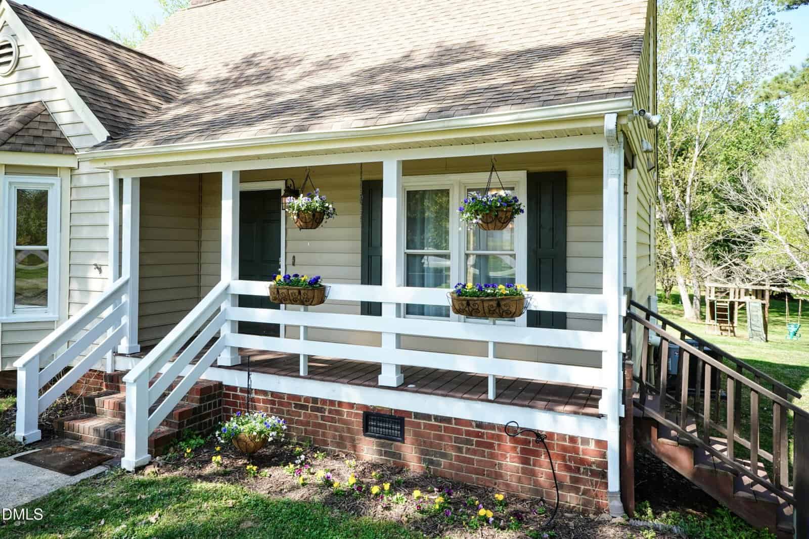 A cozy house with a brick foundation, tan siding, a front porch with white railing, and hanging flower baskets. Steps lead to the porch, and there is a grassy yard with trees in the background.