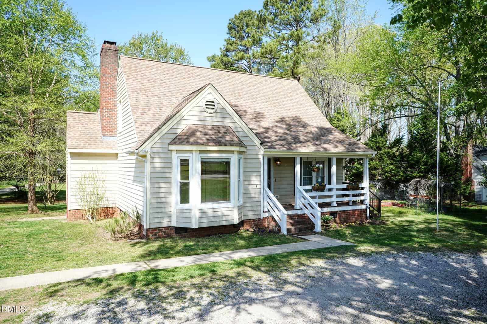 A light-colored house with a steep gabled roof, brick chimney, bay window, and a small covered front porch sits on a grassy lot with trees in the background. A gravel driveway runs in front of the house.