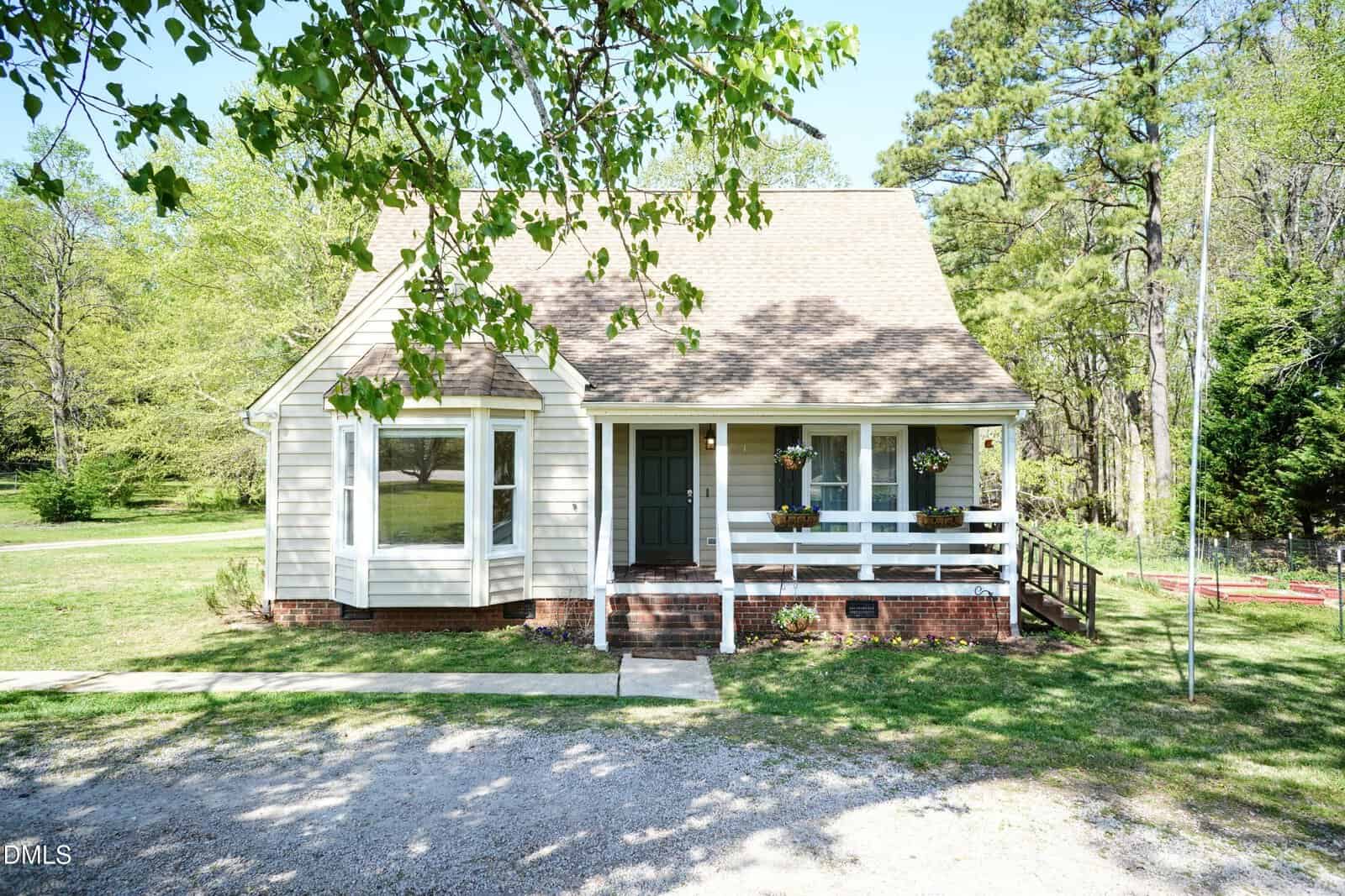 A beige house with a steep roof, front porch, and brick foundation is surrounded by trees and grass. Flower boxes hang on the porch railing, and a gravel driveway runs in front of the house.