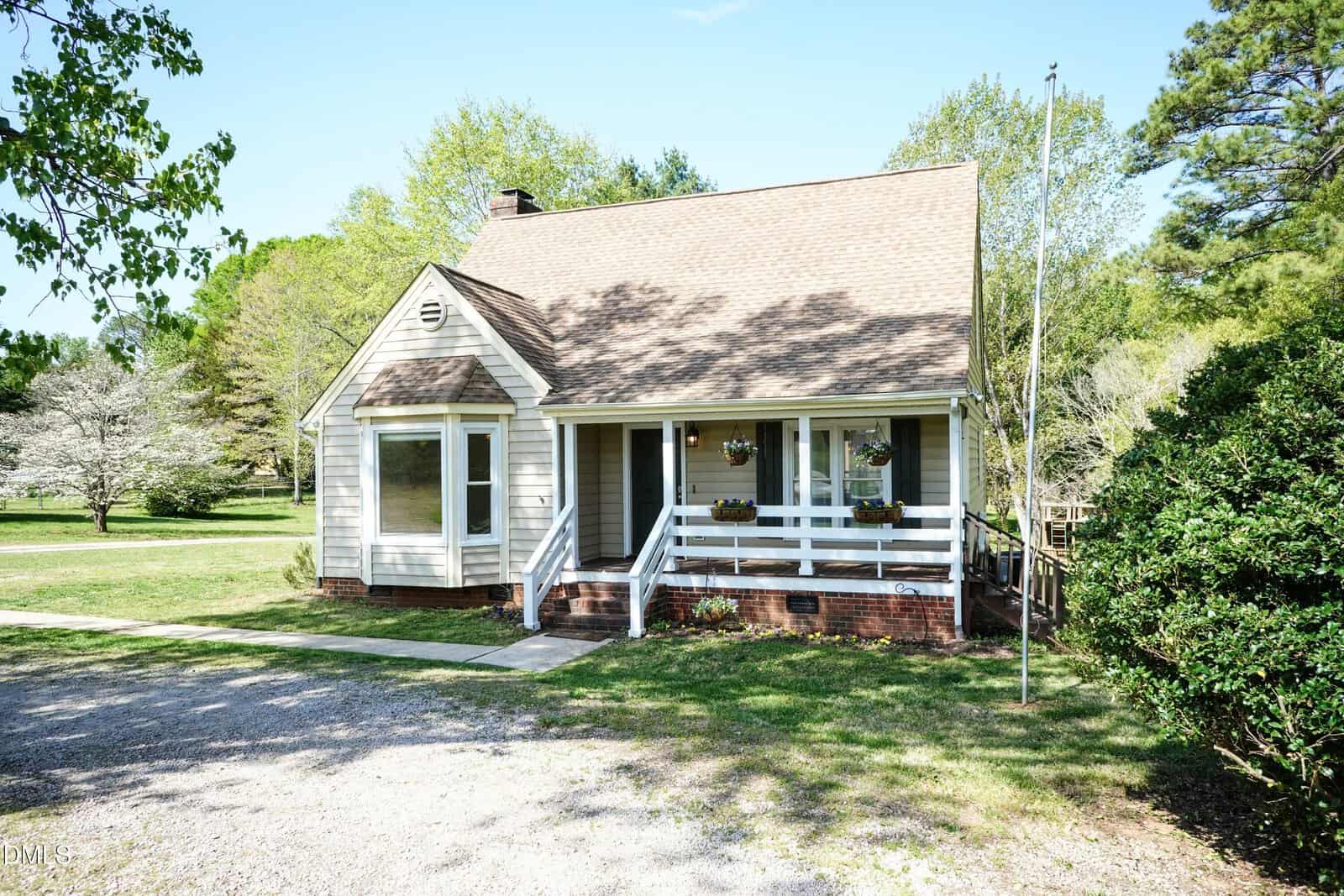 A small, light-colored house with a steep roof, front porch, and bay window sits on a grassy lot with trees in the background and a gravel driveway in the foreground. A flagpole stands near the porch.