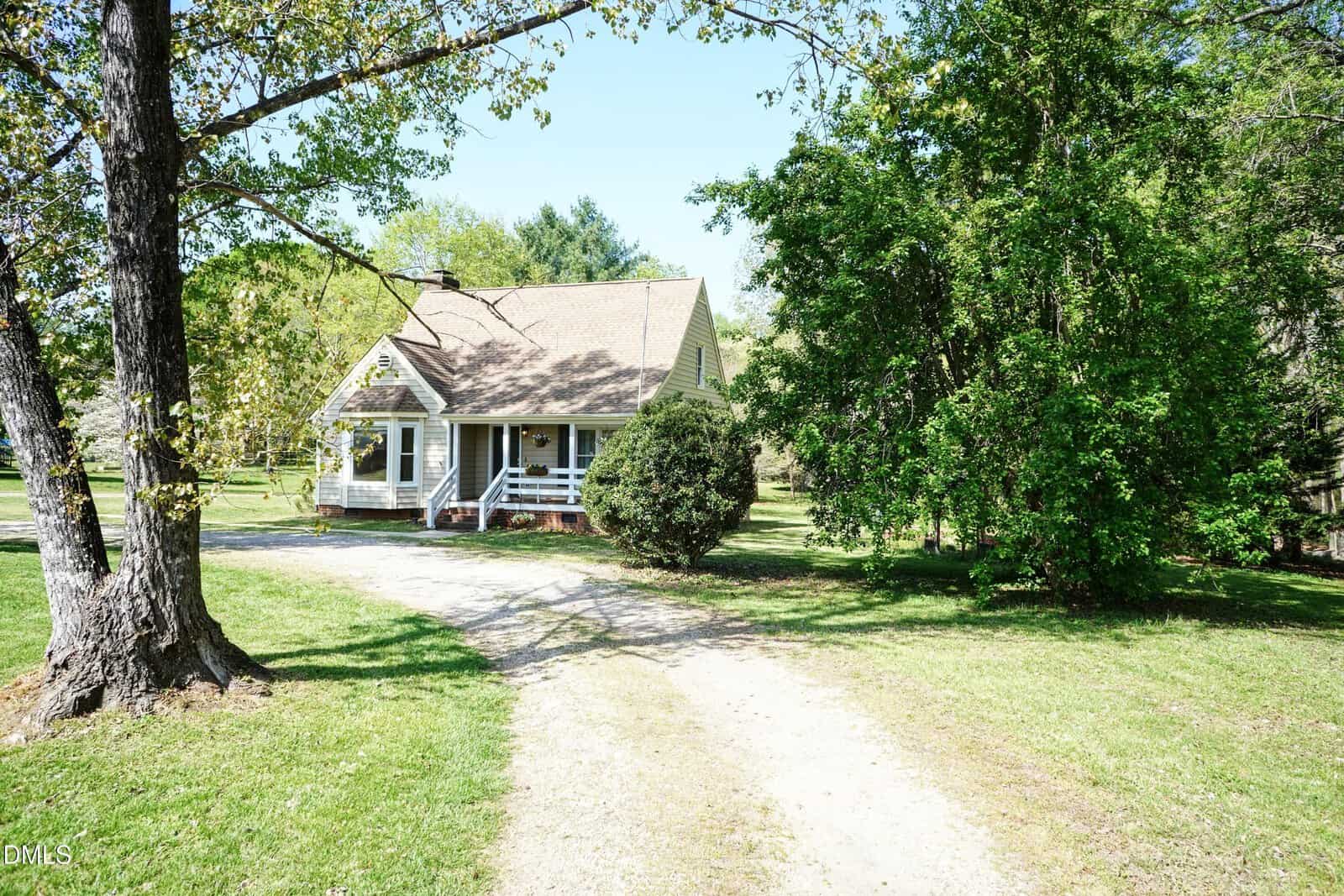 A small house with a covered front porch sits at the end of a curved gravel driveway, surrounded by green grass, trees, and shrubs under a clear blue sky.