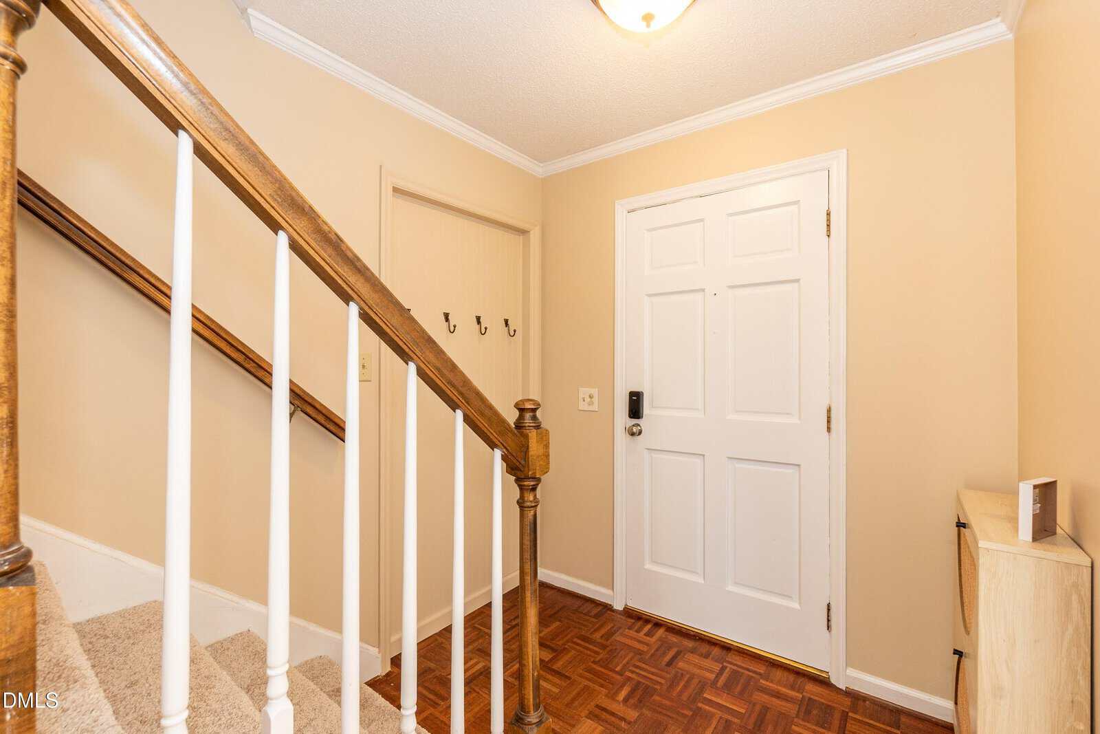 A beige entryway with a white front door, a staircase with a wooden handrail and white spindles, parquet flooring, wall hooks, and a small cabinet. Ceiling light above.