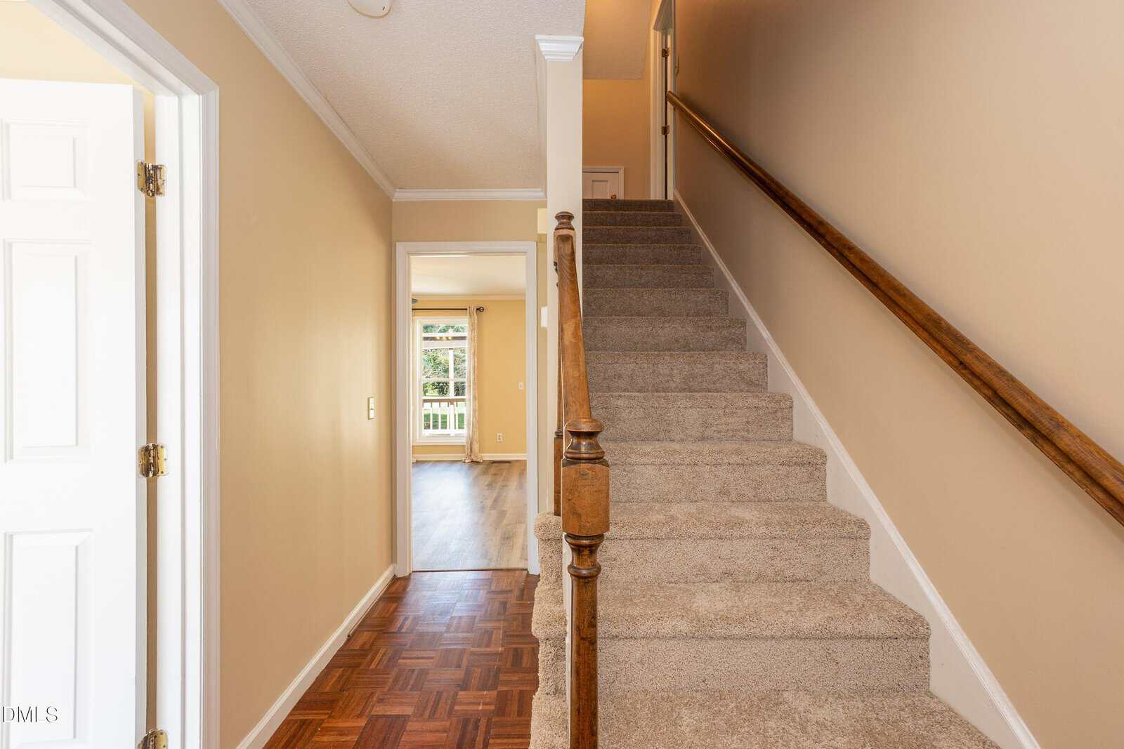 A hallway with beige walls, carpeted stairs with a wooden handrail on the right, and wooden flooring leading to a bright room with large windows in the background.