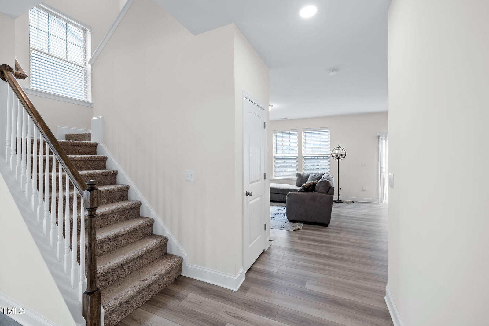 Bright, modern home interior with carpeted staircase on the left, light wood flooring, white walls, and an open living area with gray sofas and large windows in the background.