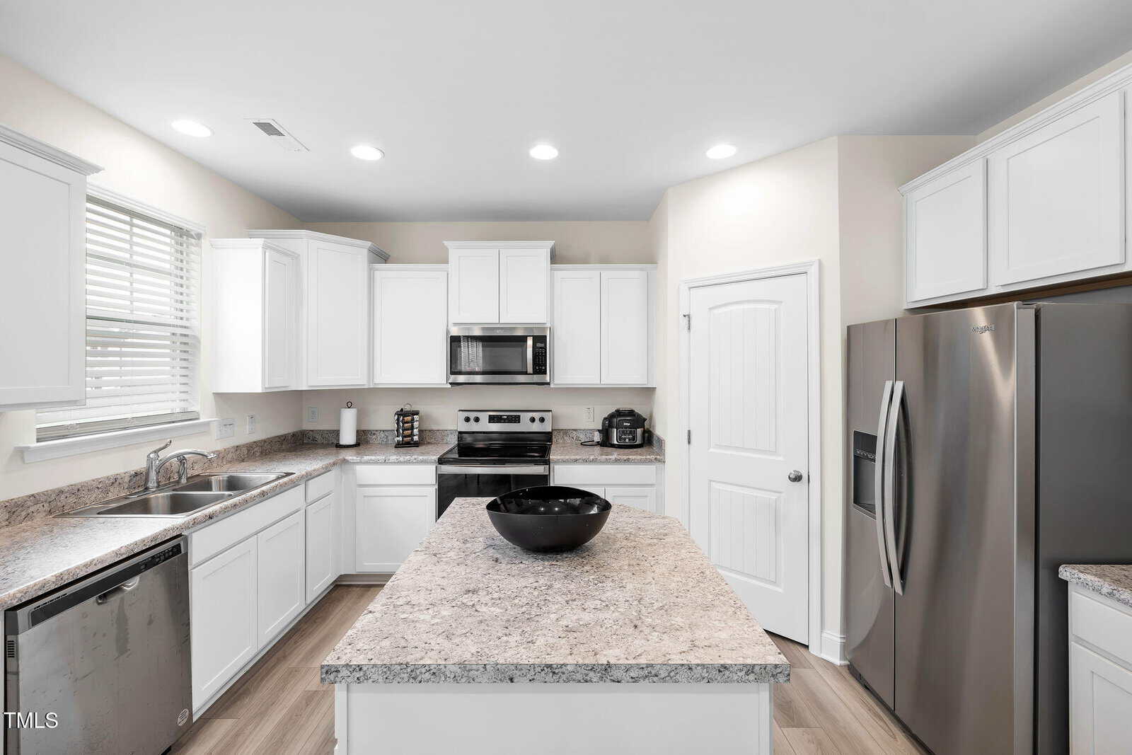 Modern kitchen with white cabinets, stainless steel appliances, a double sink, and a large central island with a black bowl. Marble-patterned counters and neutral walls create a bright, clean space.